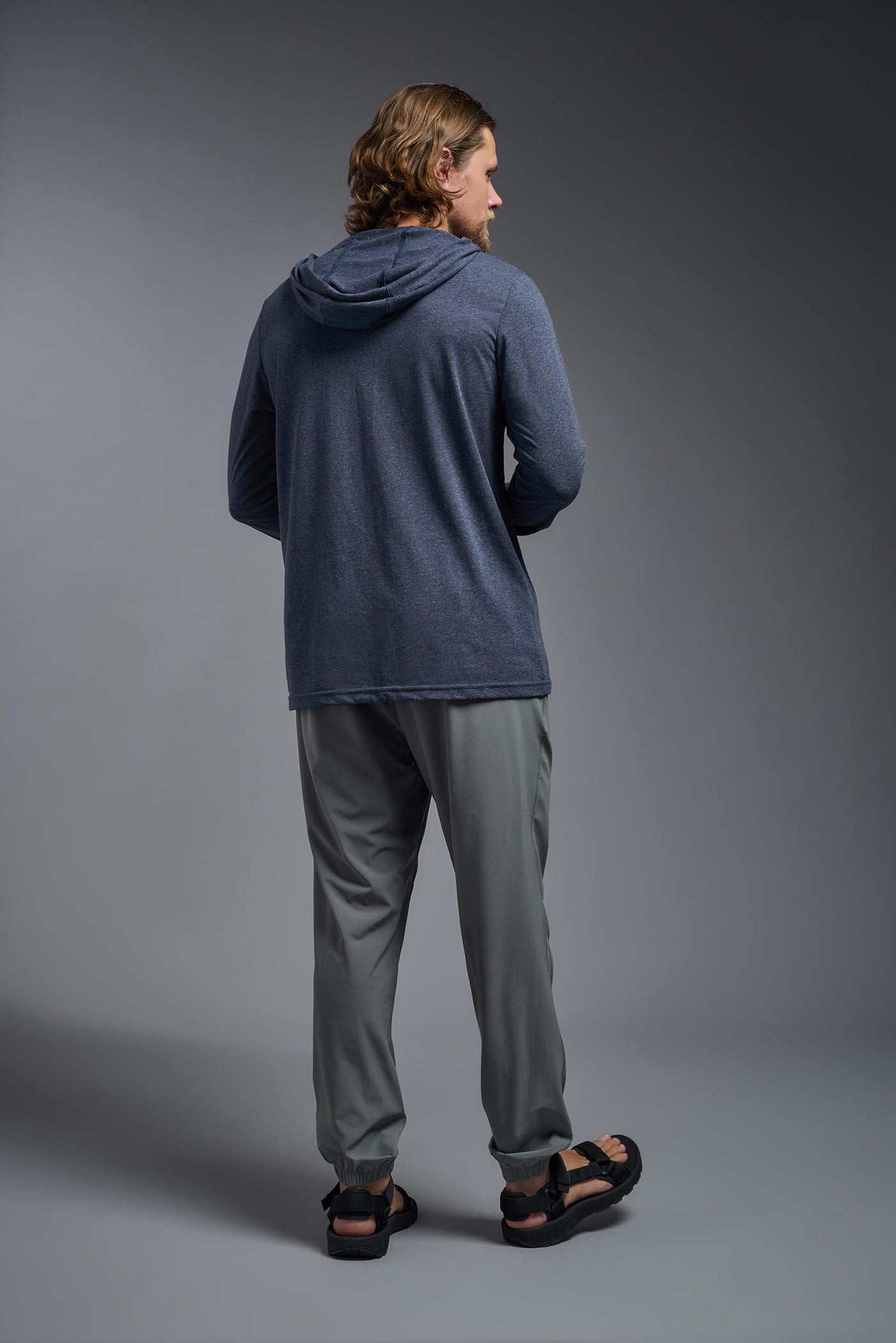 A blonde male model with a beard on a gray background, wearing the men's Heather Hoodie Shirt in the color dark heather navy, made out of 88% polyester and 22% treated cotton, showing the relaxed fit from a back view. This hoodie shirt is comfortable and made in the USA. #dark-heather-navy