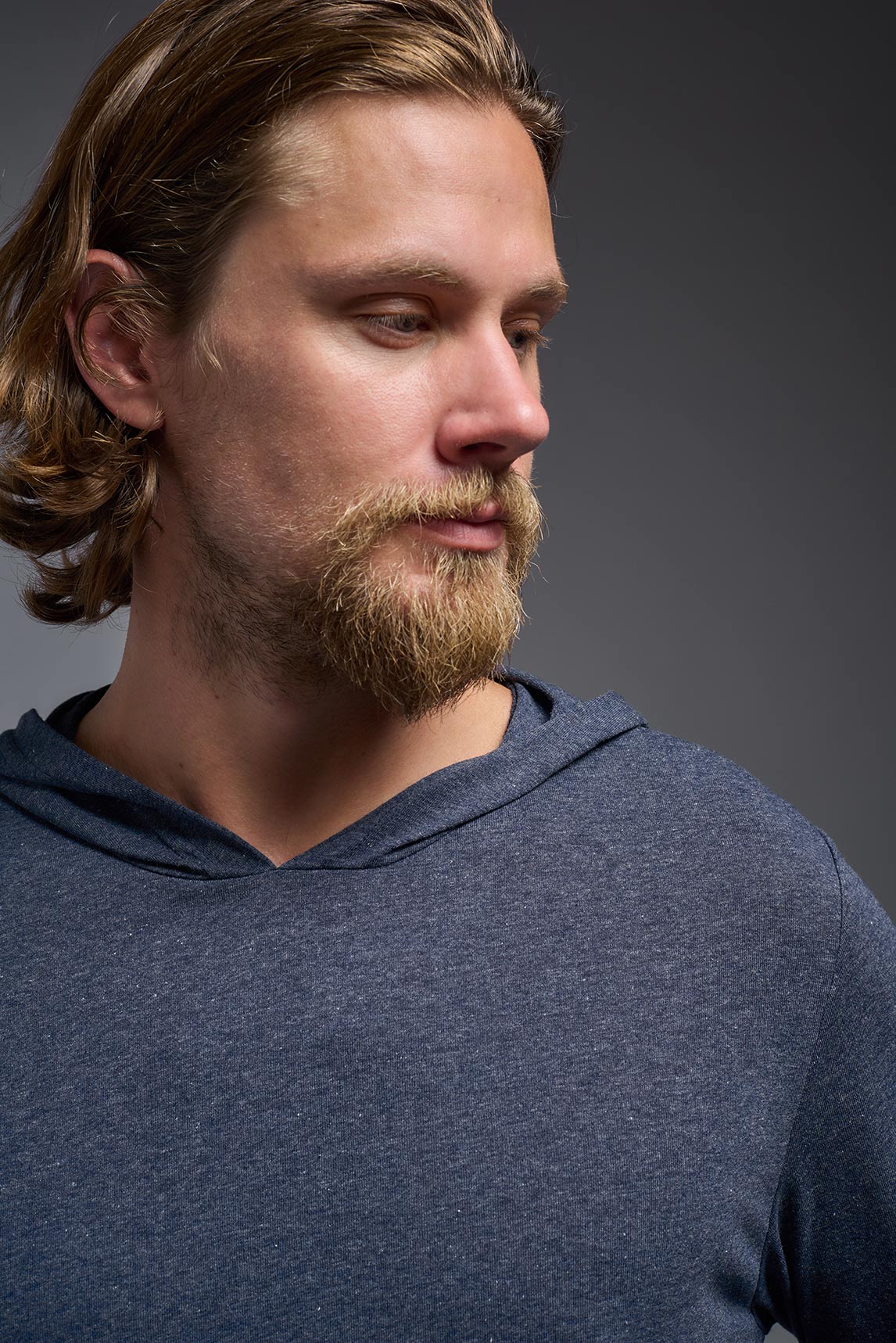 A blonde male model with a beard on a gray background, wearing the men's Heather Hoodie Shirt in the color dark heather navy, made out of 88% polyester and 22% treated cotton, showing the relaxed fit from a front closeup collar view. This hoodie shirt is comfortable and made in the USA. #dark-heather-navy