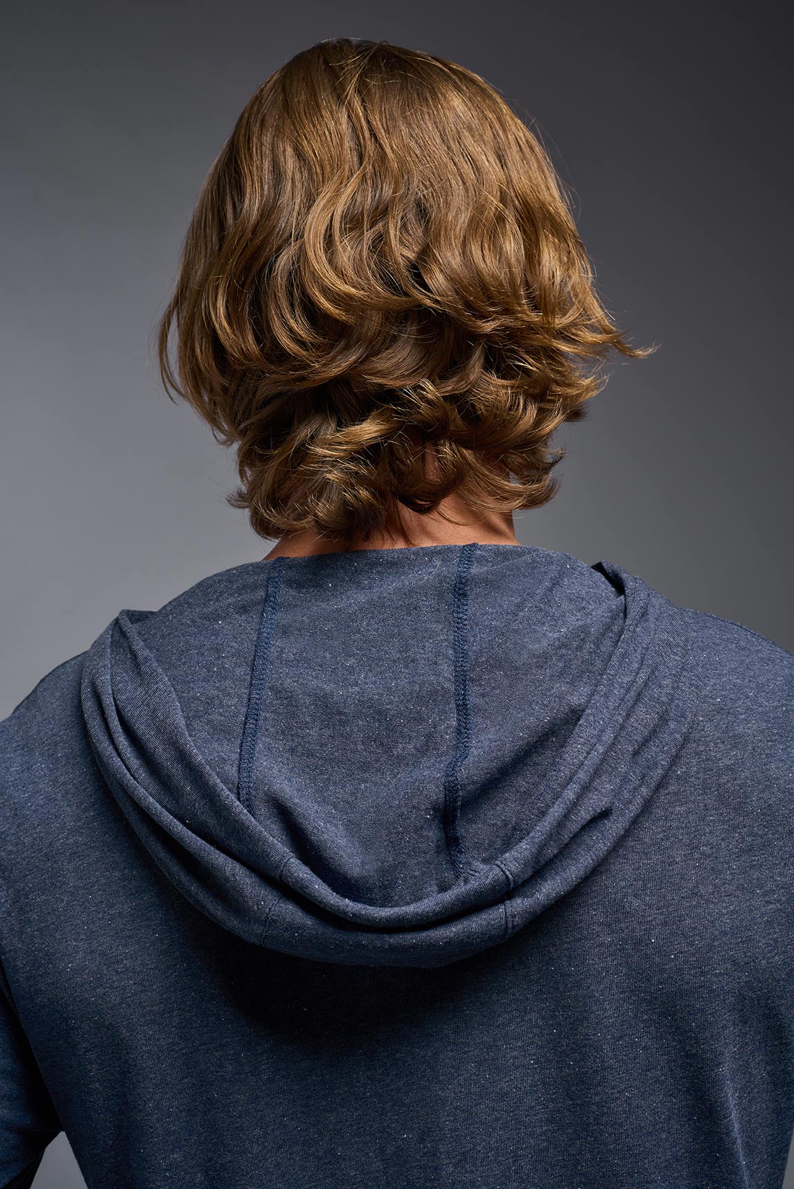 A blonde male model with a beard on a gray background, wearing the men's Heather Hoodie Shirt in the color dark heather navy, made out of 88% polyester and 22% treated cotton, showing the relaxed fit from a back closeup hoodie view. This hoodie shirt is comfortable and made in the USA. #dark-heather-navy