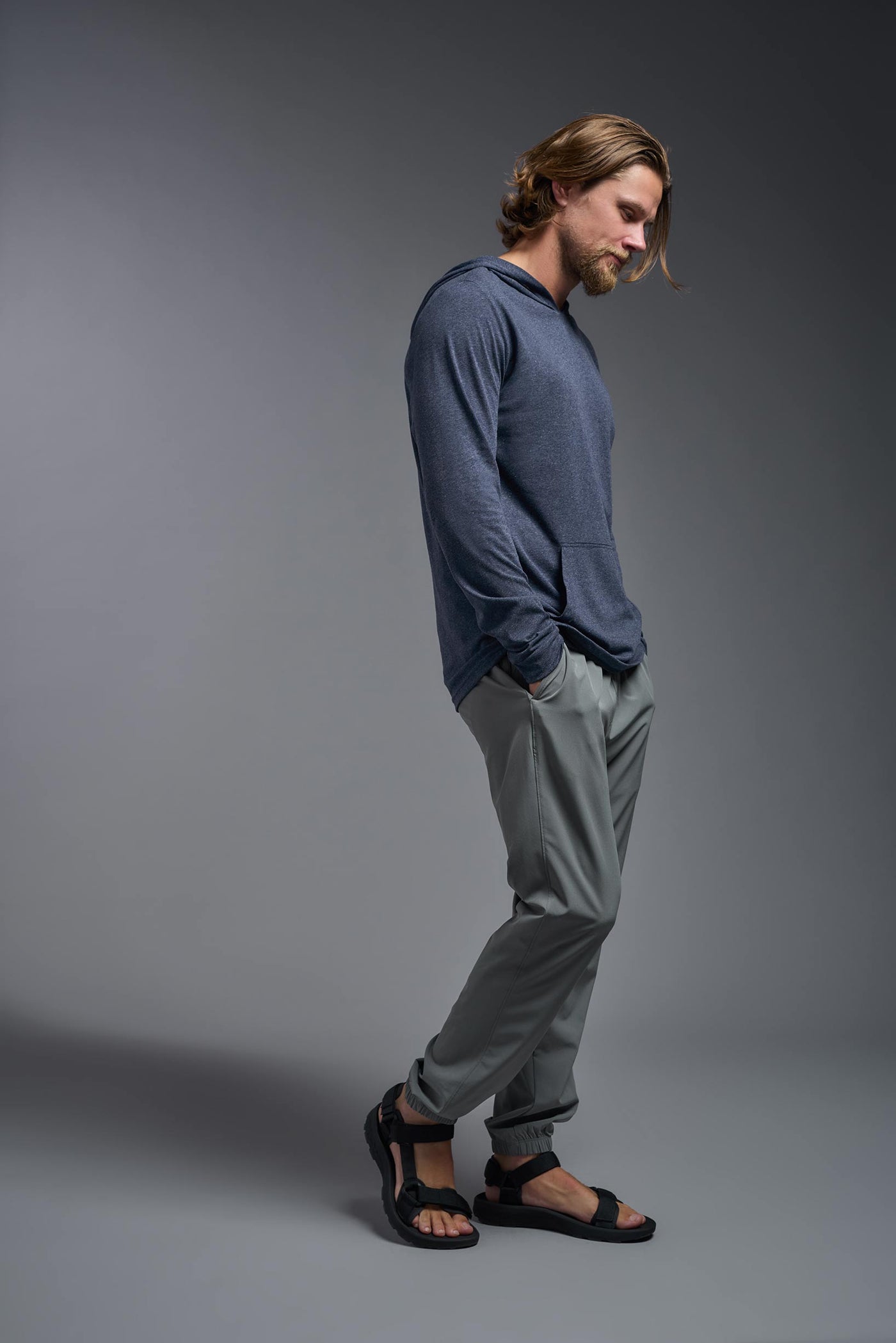 A blonde male model with a beard on a gray background, wearing the men's Heather Hoodie Shirt in the color dark heather navy, made out of 88% polyester and 22% treated cotton, showing the relaxed fit from a side view. This hoodie shirt is comfortable and made in the USA. #dark-heather-navy