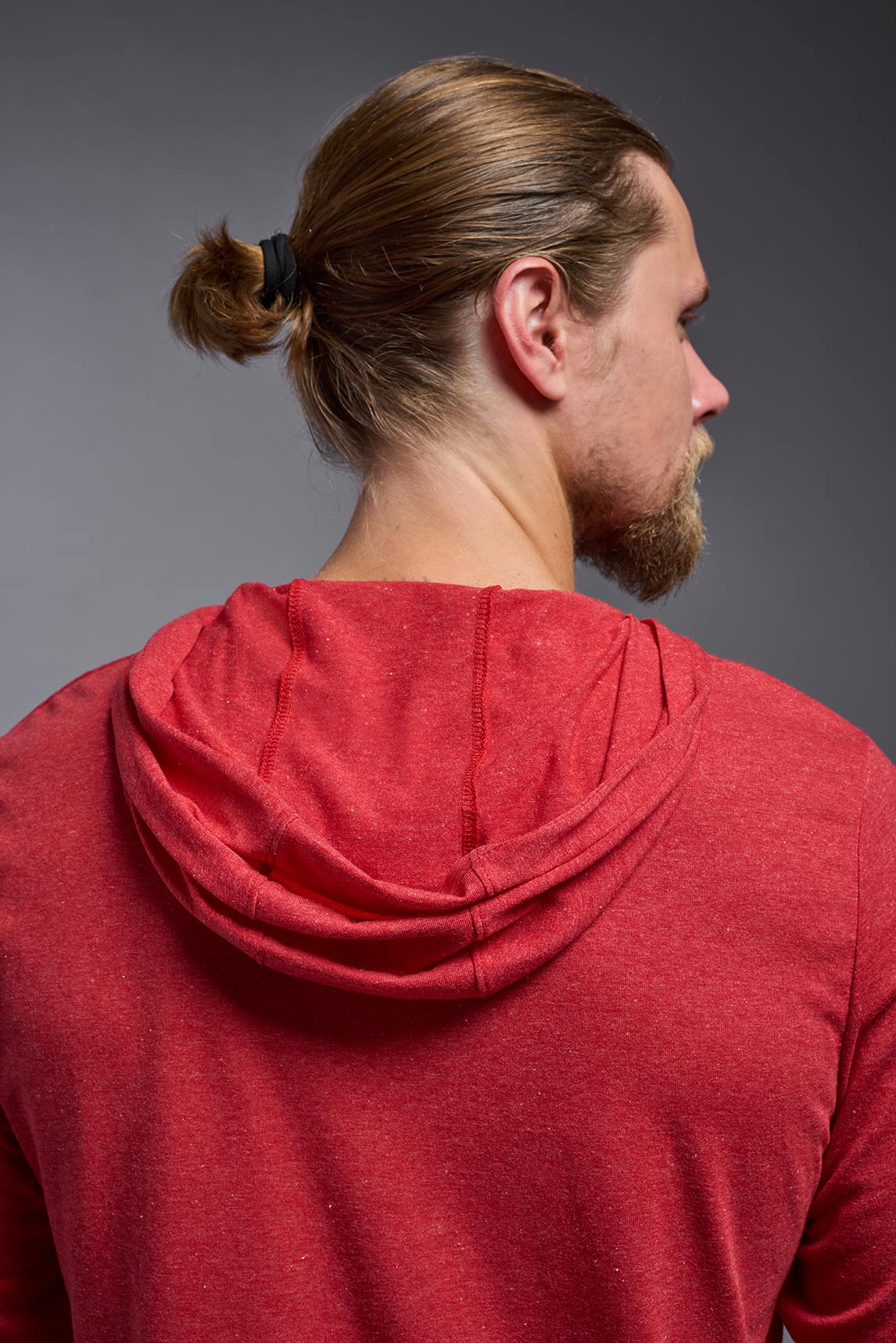 A blonde male model with a beard on a gray background, wearing the men's Heather Hoodie Shirt in the color dark heather red, made out of 88% polyester and 22% treated cotton, showing the relaxed fit from a back closeup hood view. This hoodie shirt is comfortable and made in the USA. #dark-heather-red