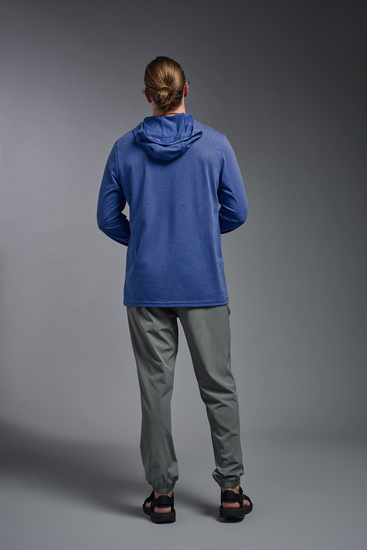 A blonde male model with a beard on a gray background, wearing the men's Heather Hoodie Shirt in the color dark heather royal, made out of 88% polyester and 22% treated cotton, showing the relaxed fit from a back view. This hoodie shirt is comfortable and made in the USA. #dark-heather-royal