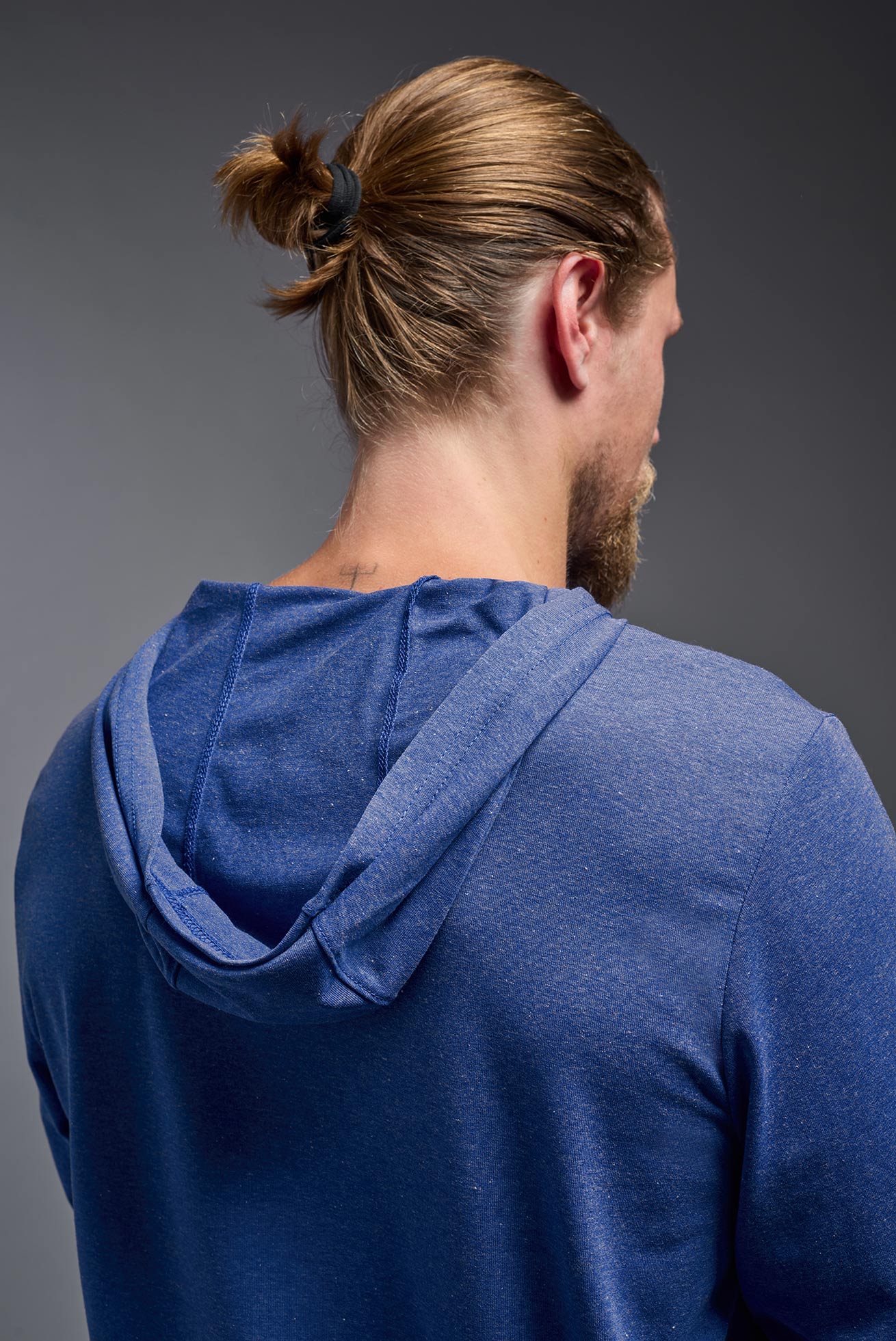 A blonde male model with a beard on a gray background, wearing the men's Heather Hoodie Shirt in the color dark heather royal, made out of 88% polyester and 22% treated cotton, showing the relaxed fit from a back closeup hoodie view. This hoodie shirt is comfortable and made in the USA. #dark-heather-royal