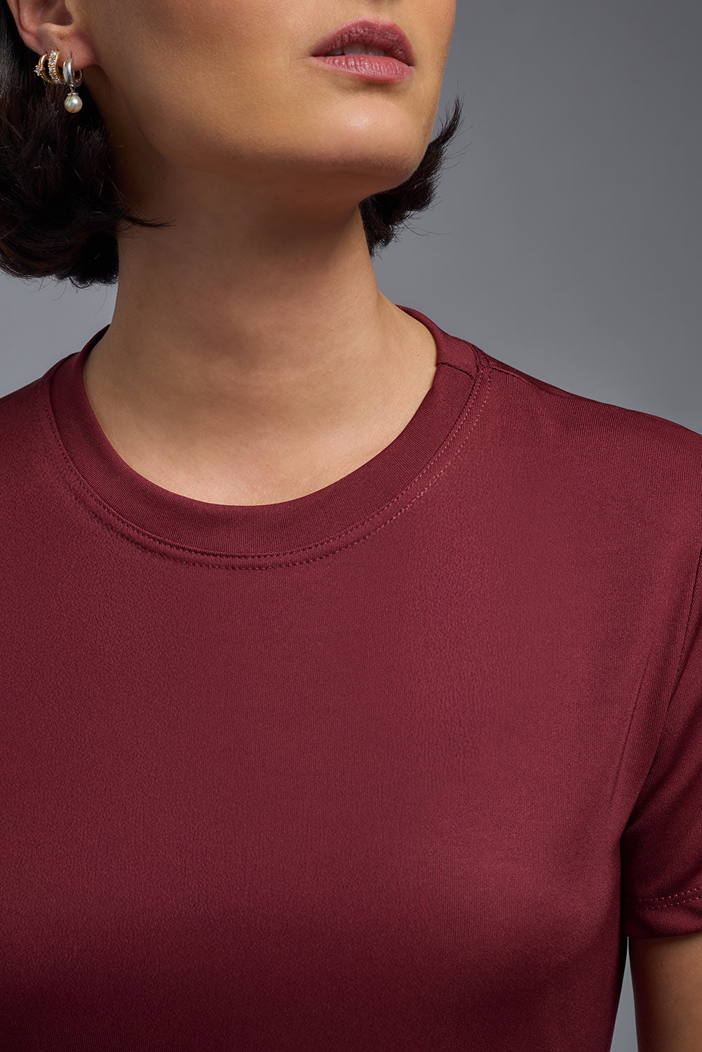 A female model with short brown hair on a gray background, wearing the Women's DriMax Crewneck Expert Tee in the color cardinal from a closeup collar front view. This shirt is made in the USA and is quick-drying and moisture-wicking. #color_cardinal