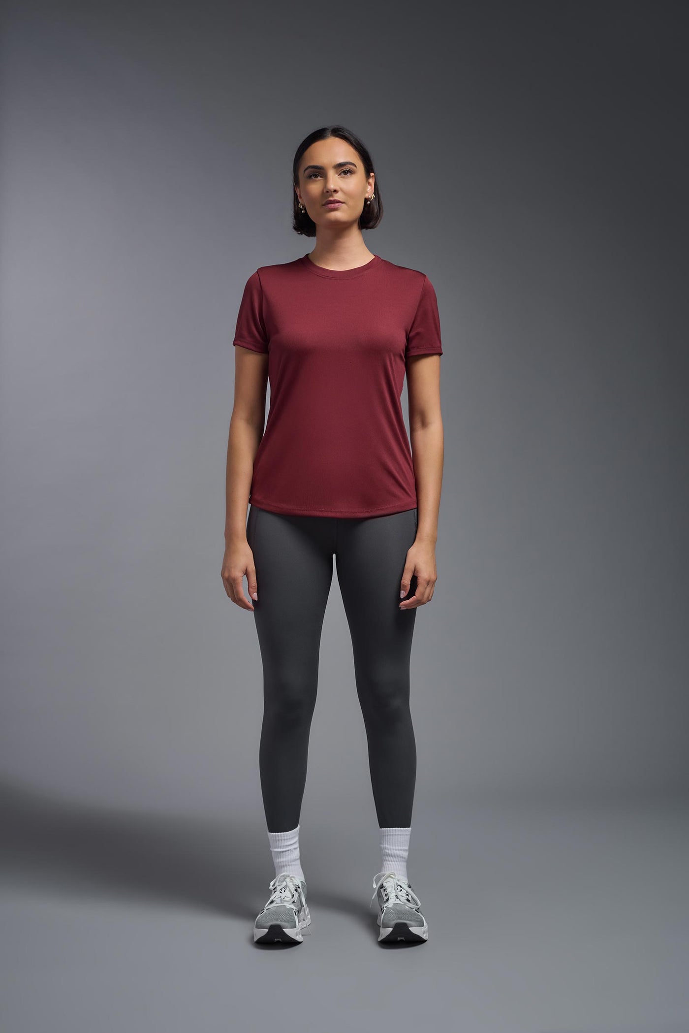 A female model with short brown hair on a gray background, wearing the Women's DriMax Crewneck Expert Tee in the color cardinal from a full body front view. This shirt is made in the USA and is quick-drying and moisture-wicking. #color_cardinal