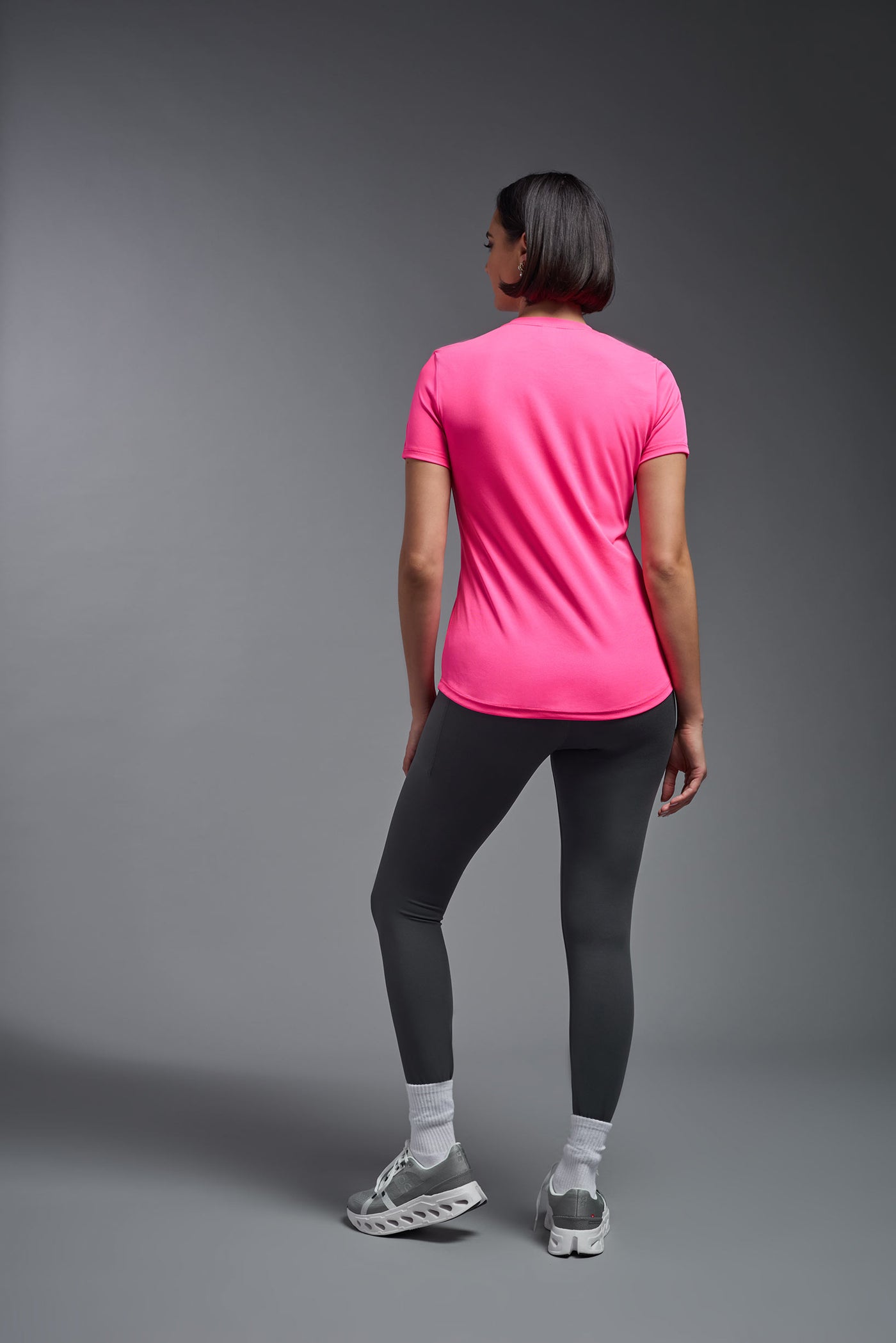 A female model with short brown hair on a gray background, wearing the Women's DriMax Crewneck Expert Tee in the color hot pink from a full body back view. This shirt is made in the USA and is quick-drying and moisture-wicking. #color_hot-pink