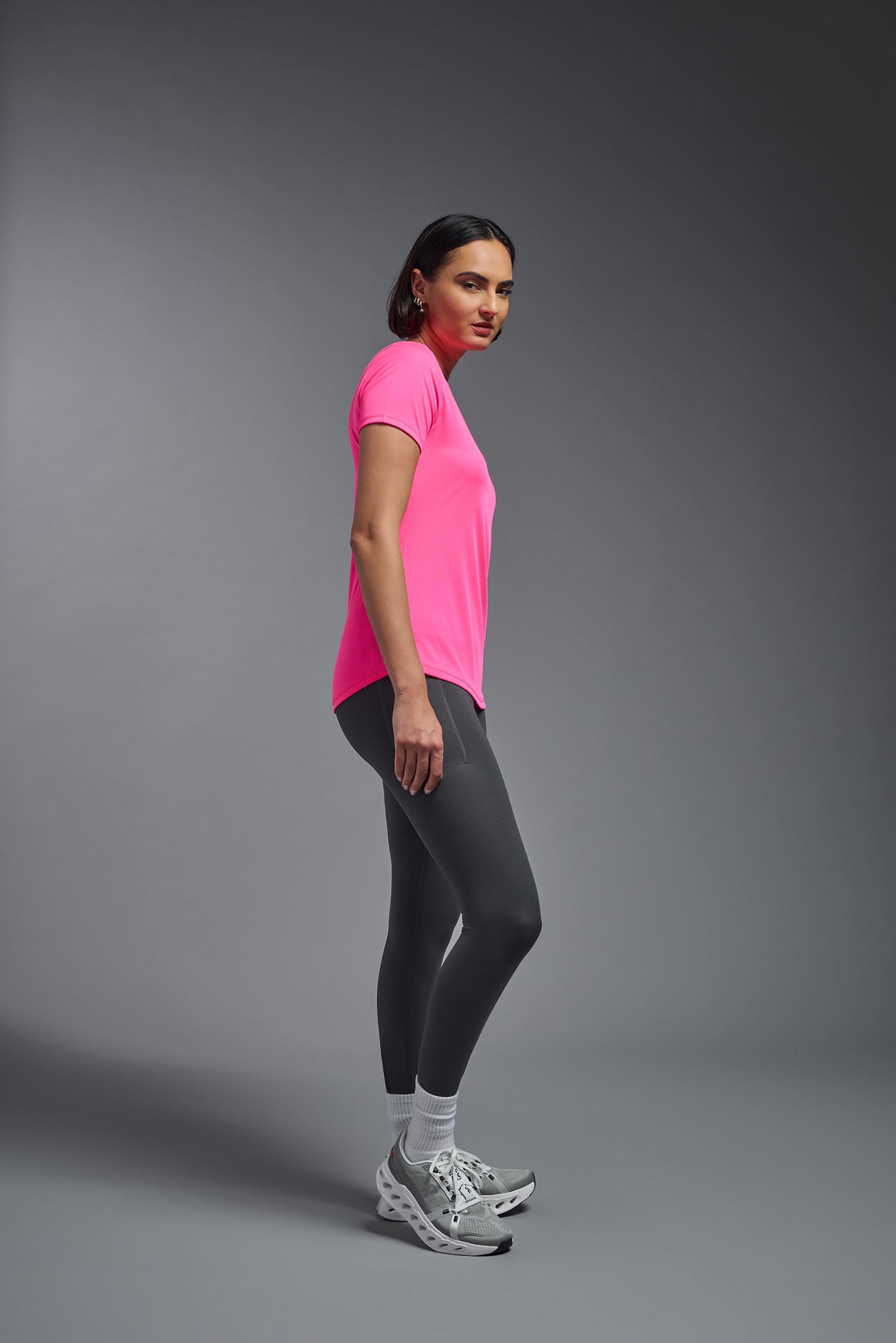 A female model with short brown hair on a gray background, wearing the Women's DriMax Crewneck Expert Tee in the color hot pink from a full body side view. This shirt is made in the USA and is quick-drying and moisture-wicking. #color_hot-pink