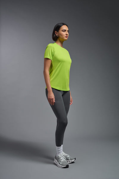 A female model with short brown hair on a gray background, wearing the Women's DriMax Crewneck Expert Tee in the color key lime from a side full body front view. This shirt is made in the USA and is quick-drying and moisture-wicking. #color_key-lime
