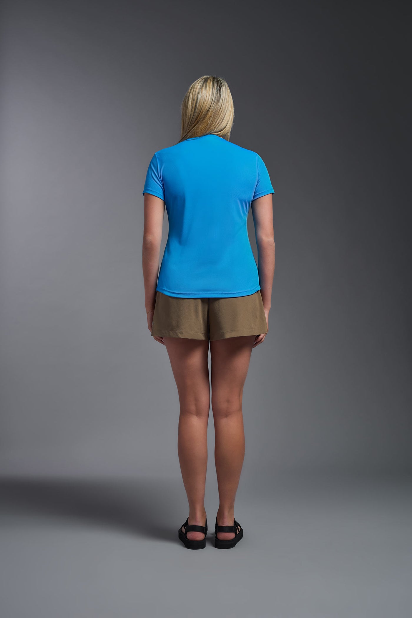 A female model with blonde straight hair on a gray background, wearing the Women's DriMax Crewneck Expert Tee in the color safety blue from a back view. This shirt is made in the USA and is quick-drying and moisture-wicking. #color_safety-blue