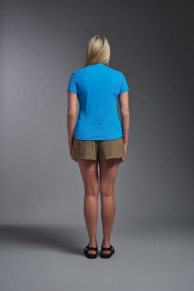 A female model with blonde straight hair on a gray background, wearing the Women's DriMax Crewneck Expert Tee in the color safety blue from a back view. This shirt is made in the USA and is quick-drying and moisture-wicking. #color_safety-blue