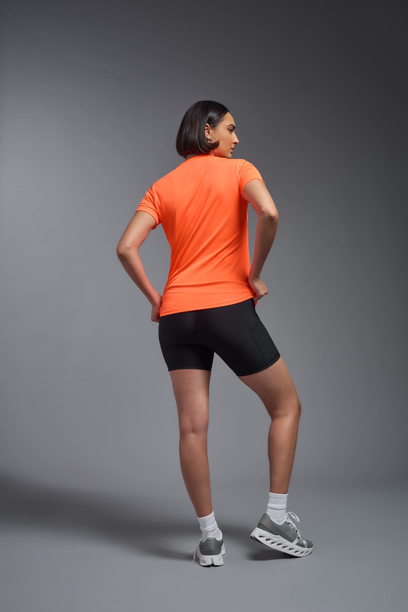 A female model with short brown hair on a gray background, wearing the Women's DriMax Crewneck Expert Tee in the color safety orange from a full body back view. This shirt is made in the USA and is quick-drying and moisture-wicking. #color_safety-orange