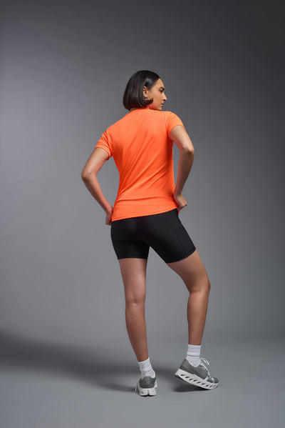 A female model with short brown hair on a gray background, wearing the Women's DriMax Crewneck Expert Tee in the color safety orange from a full body back view. This shirt is made in the USA and is quick-drying and moisture-wicking. #color_safety-orange