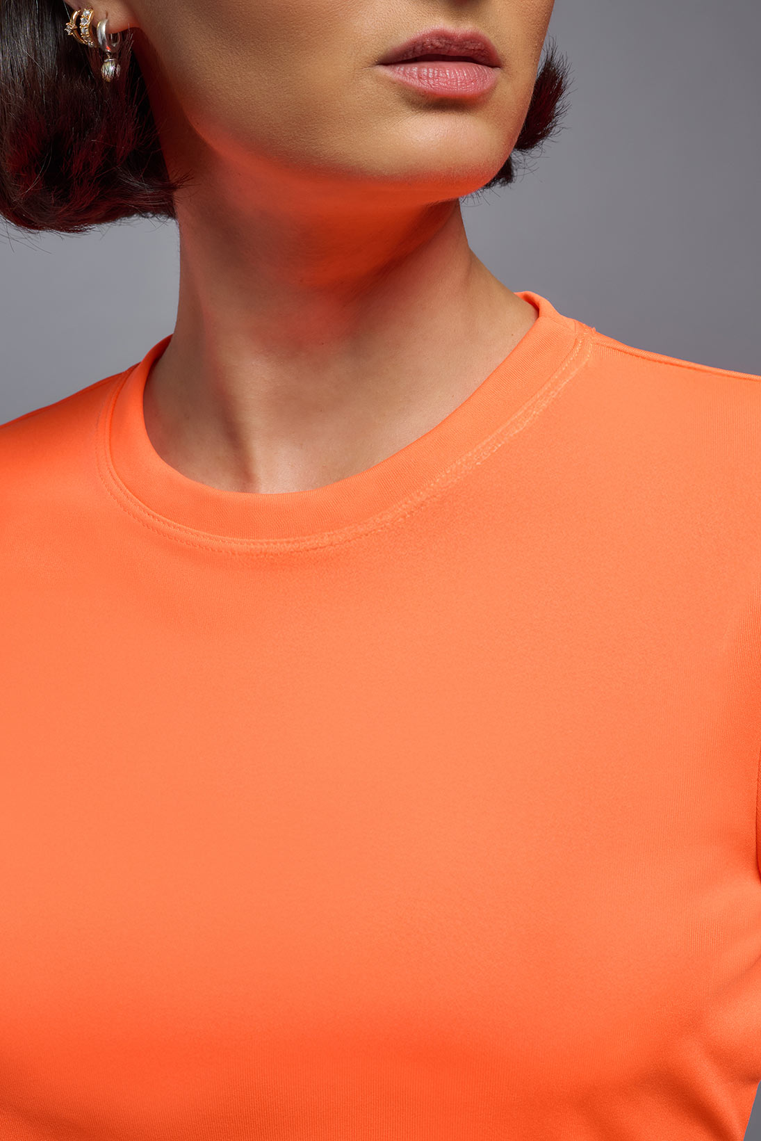 A female model with short brown hair on a gray background, wearing the Women's DriMax Crewneck Expert Tee in the color safety orange from a closeup collar front view. This shirt is made in the USA and is quick-drying and moisture-wicking. #color_safety-orange