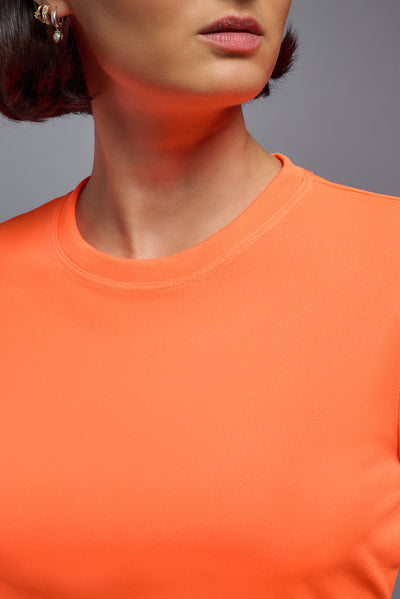 A female model with short brown hair on a gray background, wearing the Women's DriMax Crewneck Expert Tee in the color safety orange from a closeup collar front view. This shirt is made in the USA and is quick-drying and moisture-wicking. #color_safety-orange