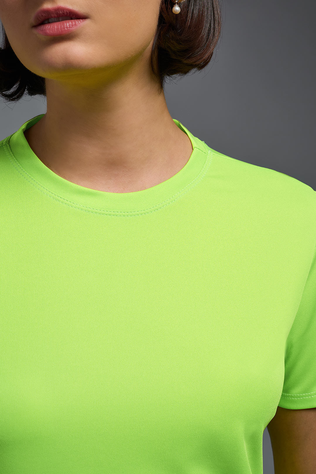 A female model with short brown hair on a gray background, wearing the Women's DriMax Crewneck Expert Tee in the color safety yellow from a closeup collar front view. This shirt is made in the USA and is quick-drying and moisture-wicking. #color_safety-yellow