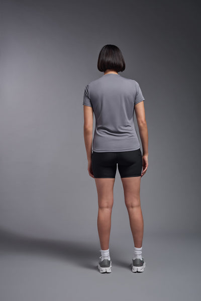 A female model with short brown hair on a gray background, wearing the Women's DriMax Crewneck Expert Tee in the color steel gray from a back view. This shirt is made in the USA and is quick-drying and moisture-wicking. #color_steel