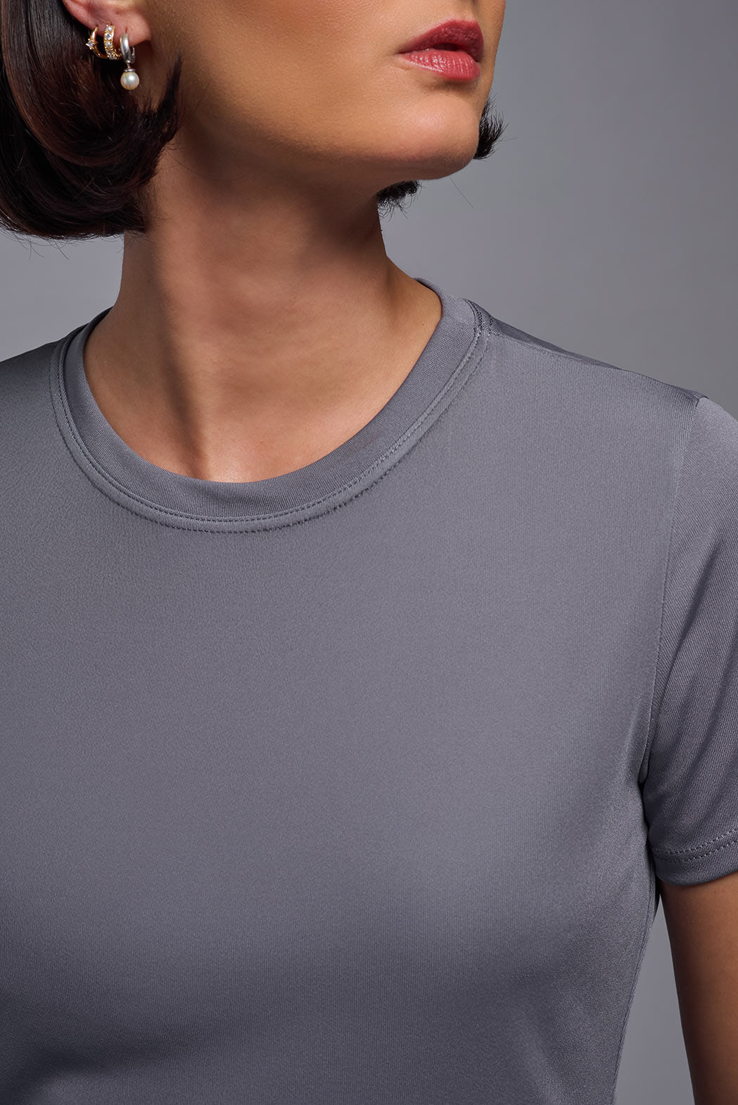 A female model with short brown hair on a gray background, wearing the Women's DriMax Crewneck Expert Tee in the color steel gray from a closeup collar front view. This shirt is made in the USA and is quick-drying and moisture-wicking. #color_steel