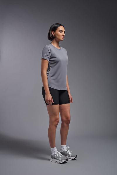 A female model with short brown hair on a gray background, wearing the Women's DriMax Crewneck Expert Tee in the color steel gray from a side view. This shirt is made in the USA and is quick-drying and moisture-wicking. #color_steel