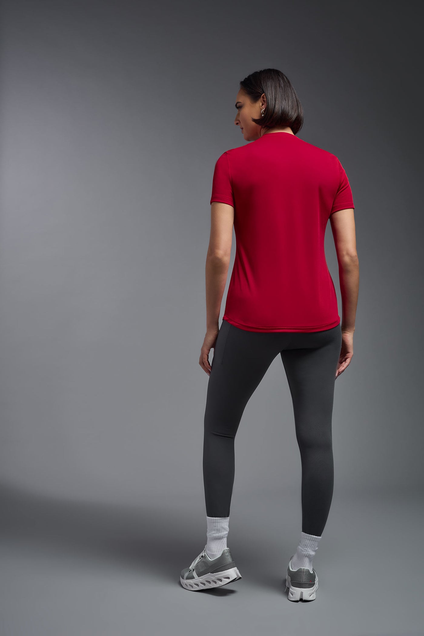 A female model with short brown hair on a gray background, wearing the Women's DriMax Crewneck Expert Tee in the color true red from a full body back view. This shirt is made in the USA and is quick-drying and moisture-wicking. #color_red