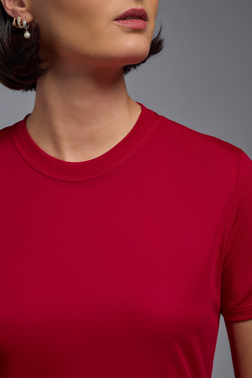 A female model with short brown hair on a gray background, wearing the Women's DriMax Crewneck Expert Tee in the color true red from a closeup collar front view. This shirt is made in the USA and is quick-drying and moisture-wicking. #color_red