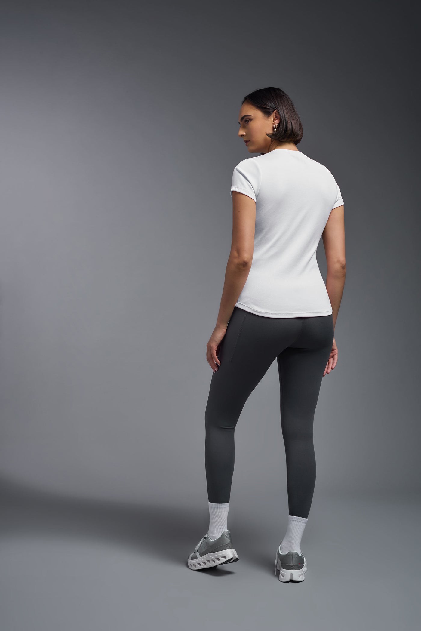 A female model with short brown hair on a gray background, wearing the Women's DriMax Crewneck Expert Tee in the color white from a full body back view. This shirt is made in the USA and is quick-drying and moisture-wicking. #color_white