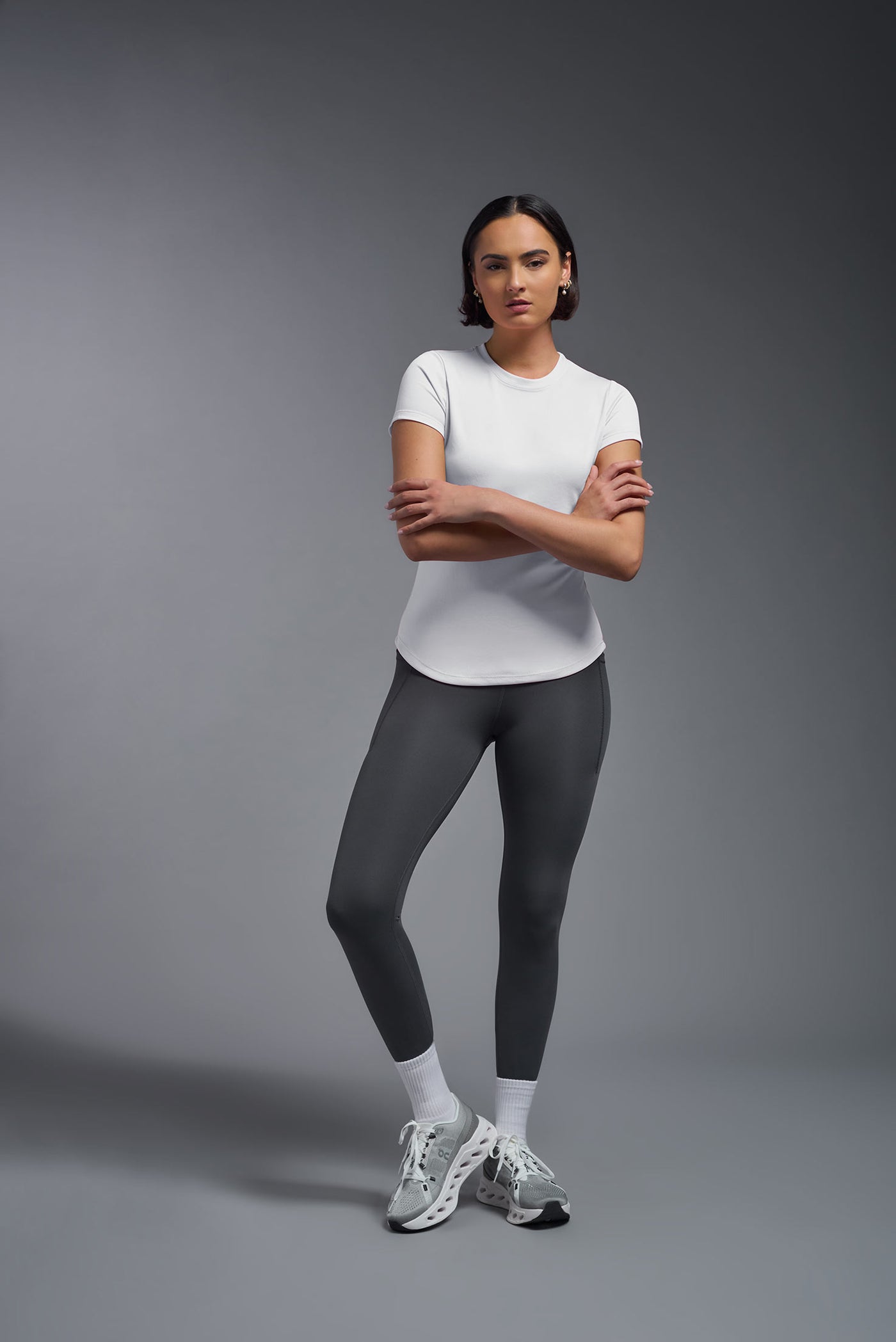 A female model with short brown hair on a gray background, wearing the Women's DriMax Crewneck Expert Tee in the color white from a full body front view. This shirt is made in the USA and is quick-drying and moisture-wicking. #color_white