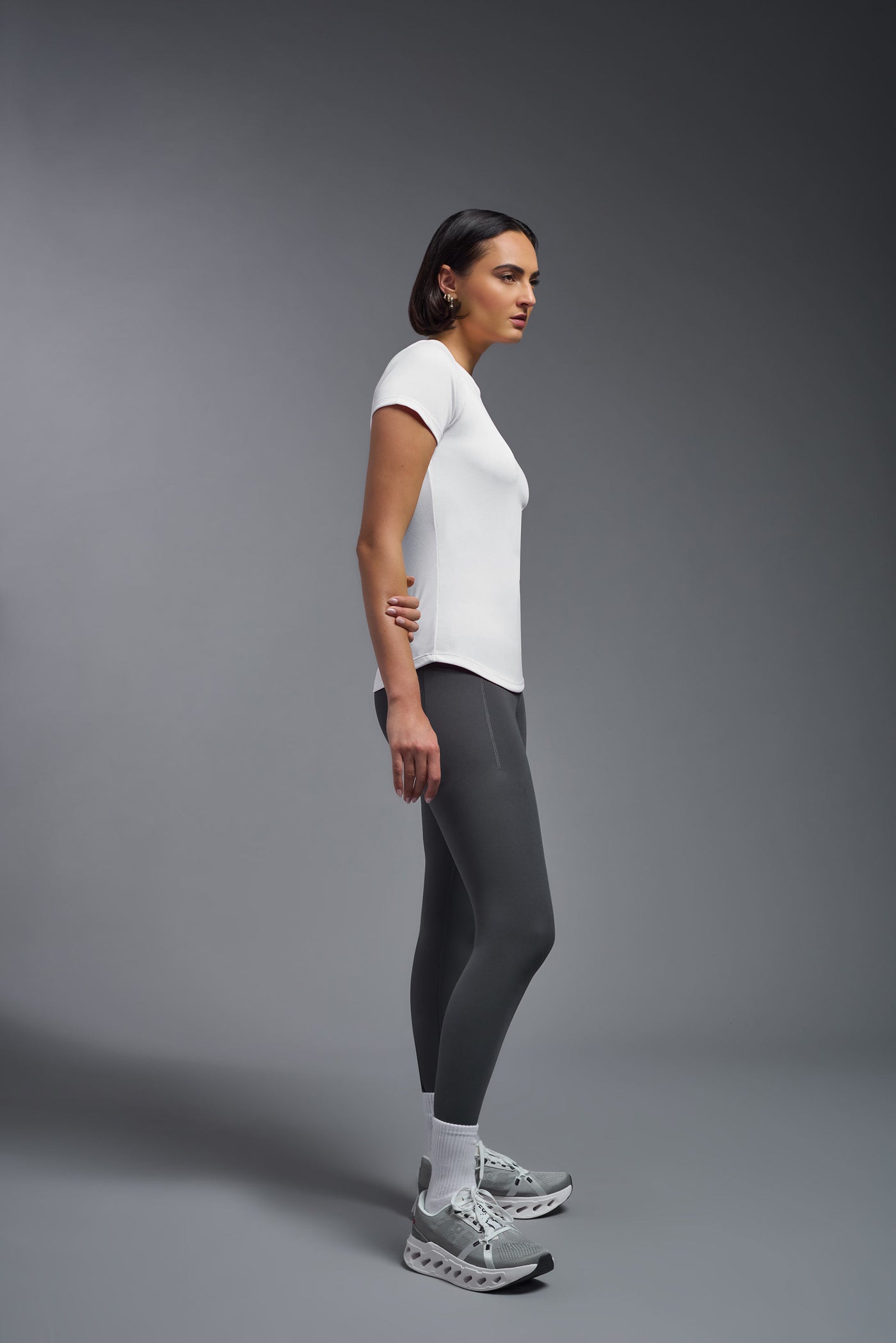A female model with short brown hair on a gray background, wearing the Women's DriMax Crewneck Expert Tee in the color white from a full body side view. This shirt is made in the USA and is quick-drying and moisture-wicking. #color_white