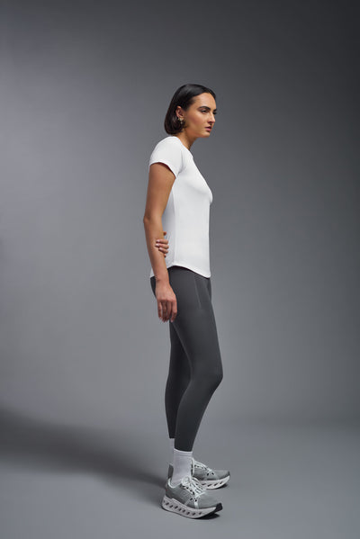 A female model with short brown hair on a gray background, wearing the Women's DriMax Crewneck Expert Tee in the color white from a full body side view. This shirt is made in the USA and is quick-drying and moisture-wicking. #color_white