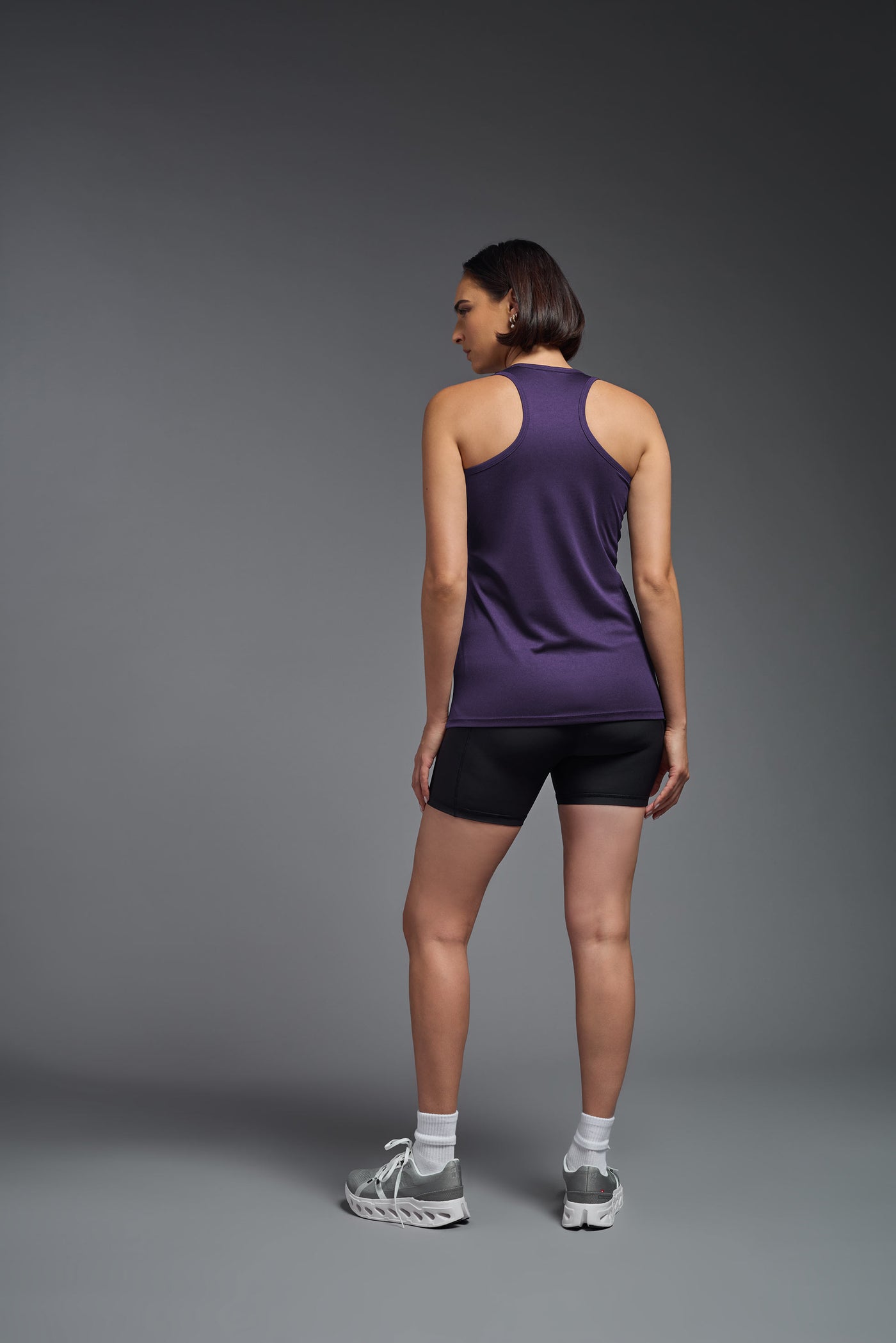 A female model with brown short hair on a gray background, wearing the Women's DrImax Endurance Racerback Tank in the color dark purple from a full body back view. This tank is made in the USA and is quick-drying and moisture-wicking. #color_dark-purple