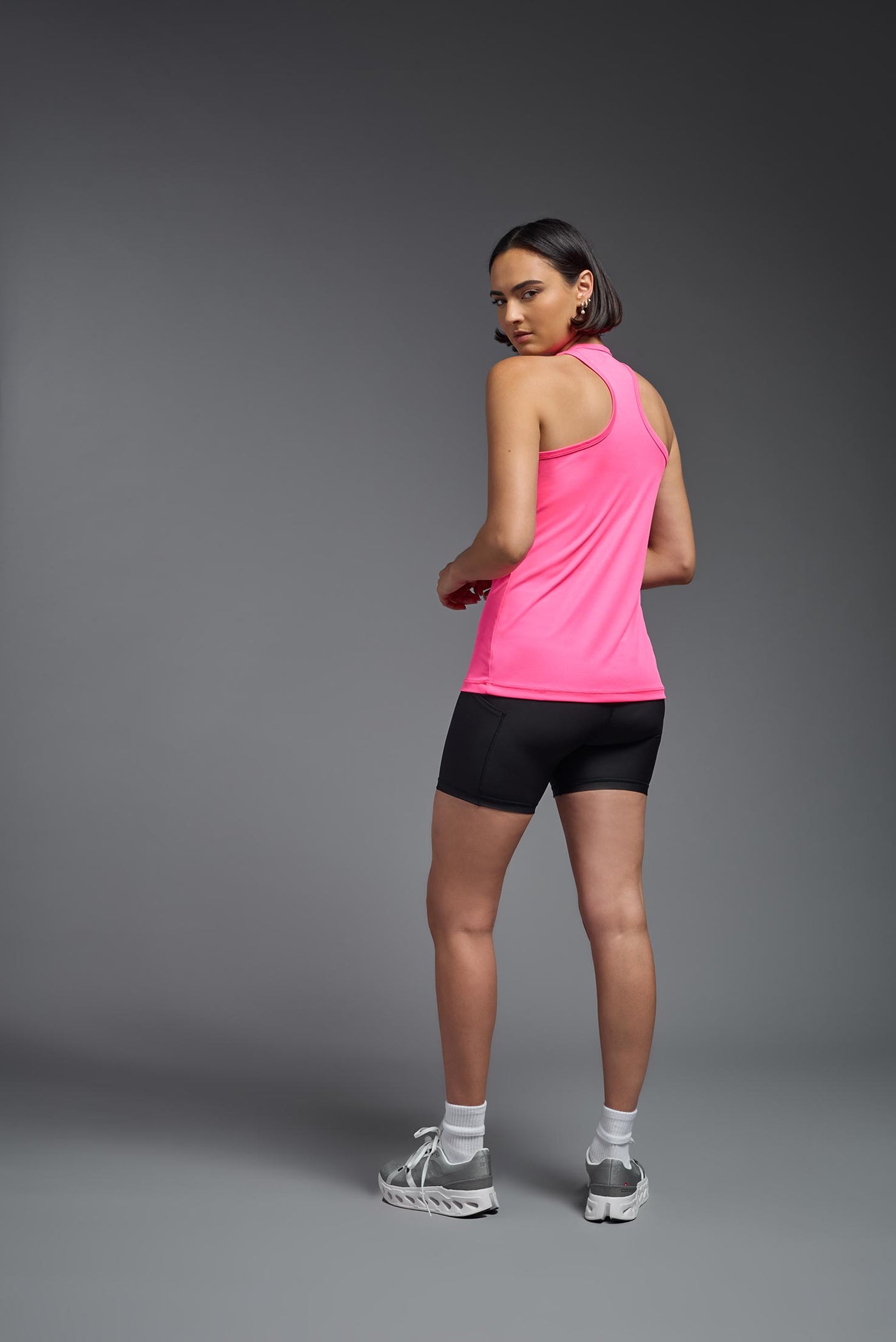 A female model with brown short hair on a gray background, wearing the Women's DrImax Endurance Racerback Tank in the color hot pink from a full body back view. This tank is made in the USA and is quick-drying and moisture-wicking. #color_hot-pink