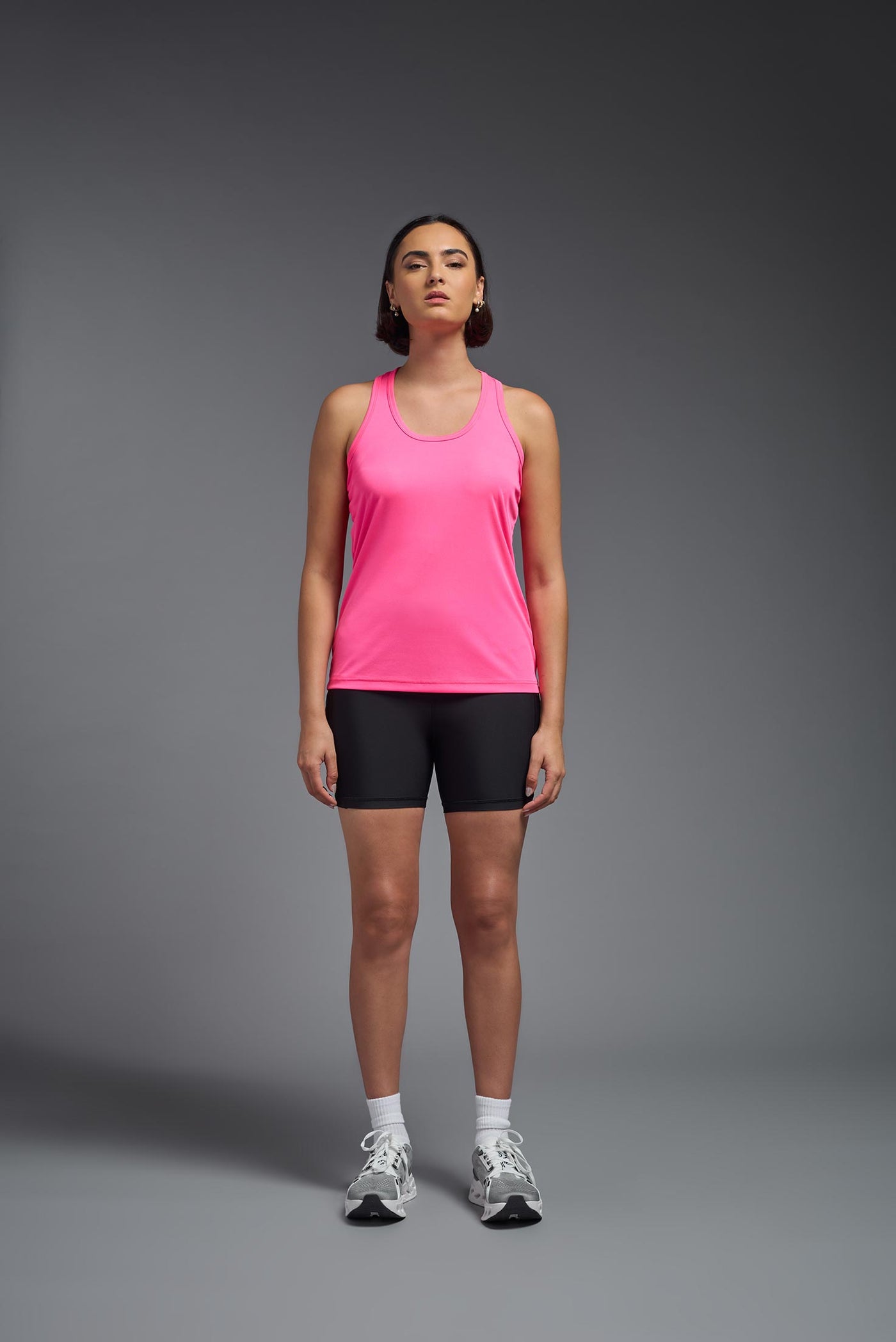 A female model with brown short hair on a gray background, wearing the Women's DrImax Endurance Racerback Tank in the color hot pink from a full body front view. This tank is made in the USA and is quick-drying and moisture-wicking. #color_hot-pink