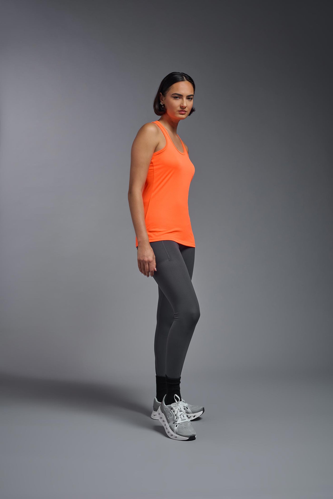 A female model with brown short hair on a gray background, wearing the Women's DrImax Endurance Racerback Tank in the color safety orange from a full body side view. This tank is made in the USA and is quick-drying and moisture-wicking. #color_safety-orange
