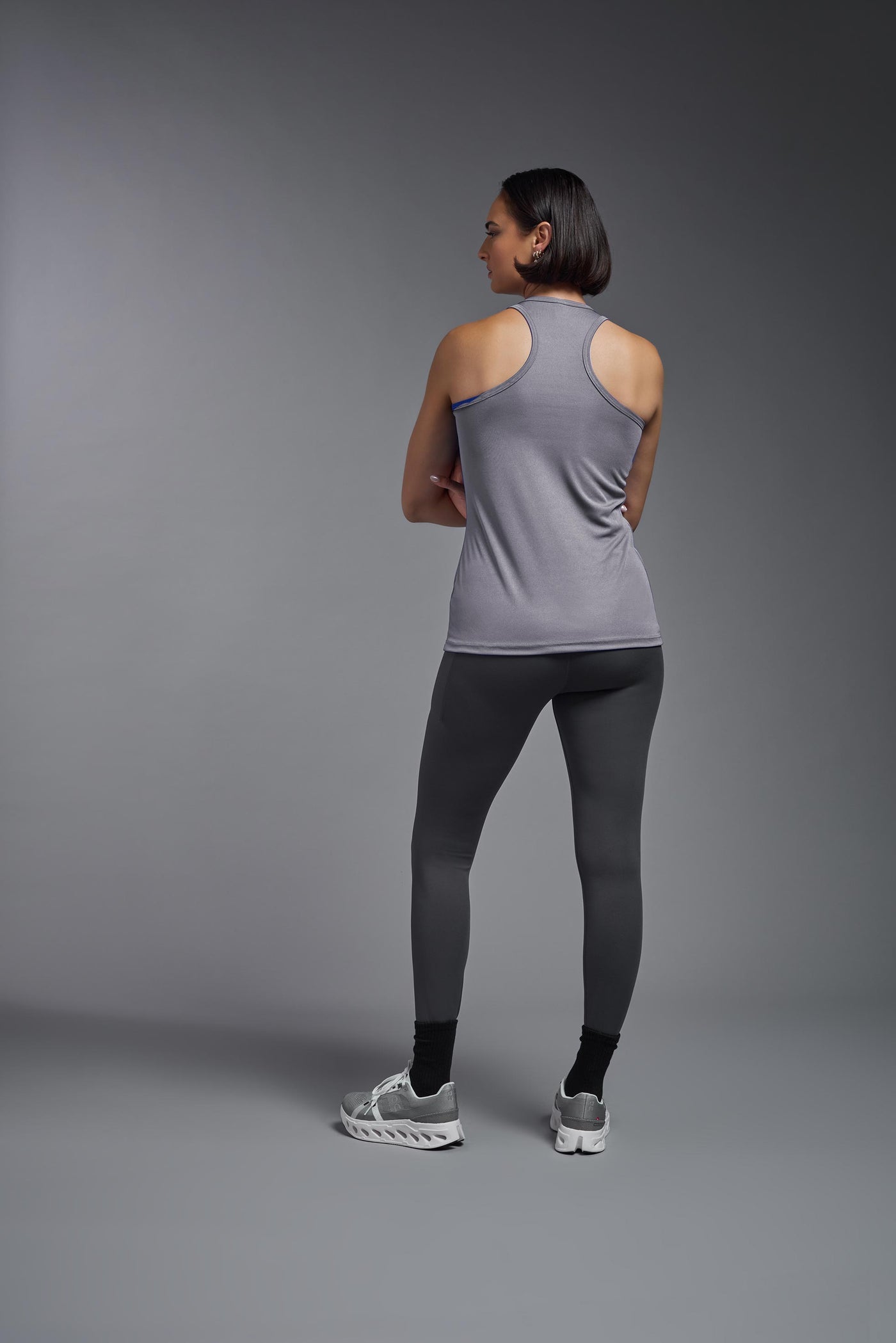 A female model with brown short hair on a gray background, wearing the Women's Drimax Endurance Racerback Tank in the color steel from a full body back view. This tank is made in the USA and is quick-drying and moisture-wicking. #color_steel