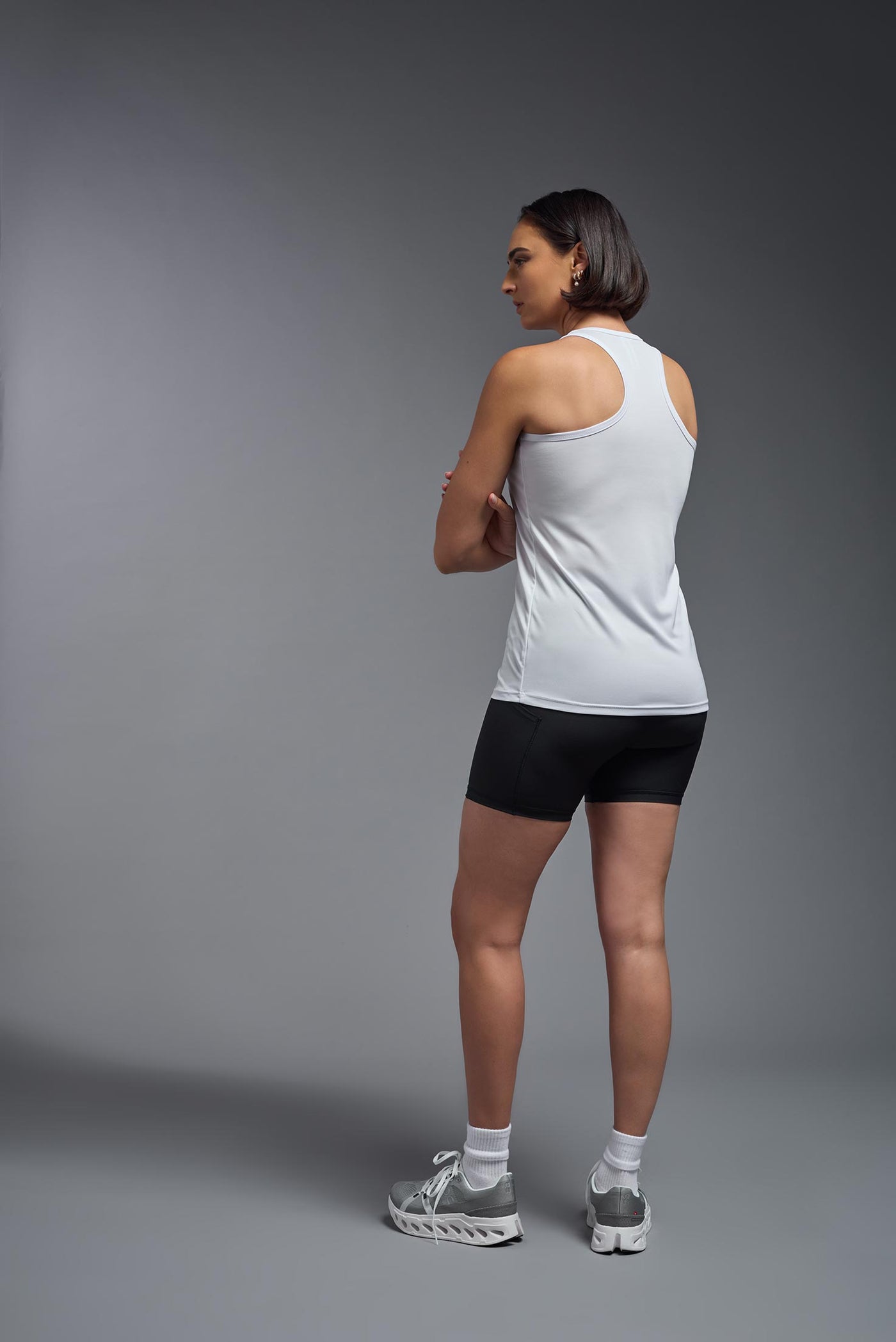 A female model with brown short hair on a gray background, wearing the Women's DrImax Endurance Racerback Tank in the color white from a full body back view. This tank is made in the USA and is quick-drying and moisture-wicking. #color_white