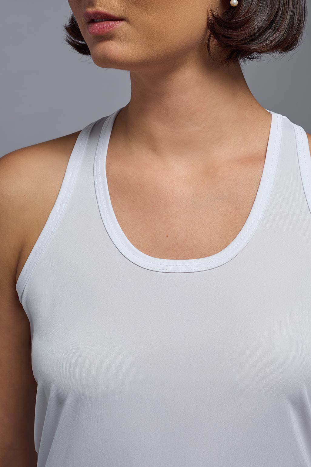 A female model with brown short hair on a gray background, wearing the Women's DrImax Endurance Racerback Tank in the color white from a front closeup collar view. This tank is made in the USA and is quick-drying and moisture-wicking. #color_white