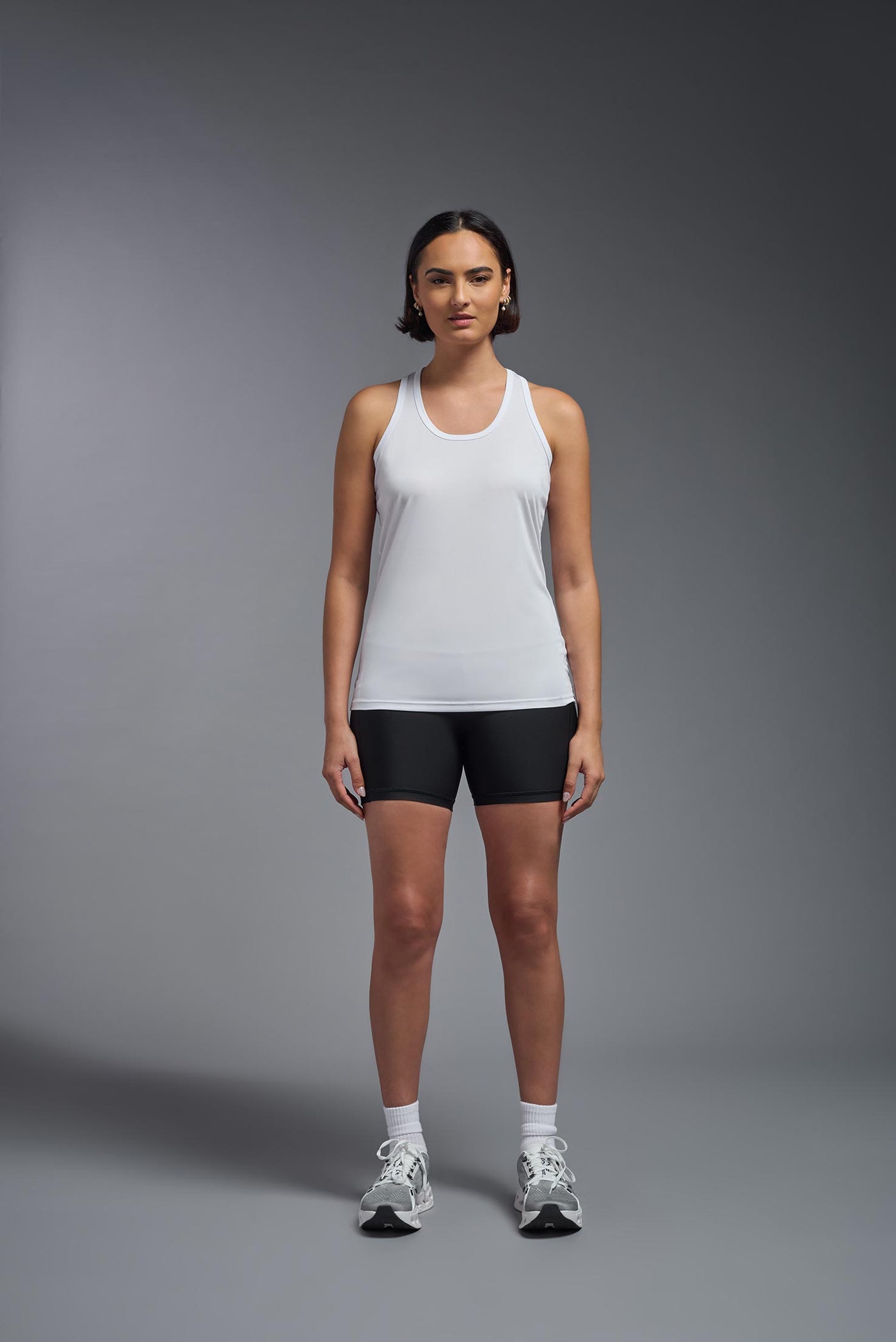 A female model with brown short hair on a gray background, wearing the Women's DrImax Endurance Racerback Tank in the color white from a full body front view. This tank is made in the USA and is quick-drying and moisture-wicking. #color_white
