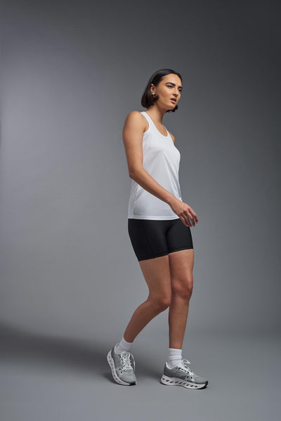 A female model with brown short hair on a gray background, wearing the Women's DrImax Endurance Racerback Tank in the color white from a full body side view. This tank is made in the USA and is quick-drying and moisture-wicking. #color_white