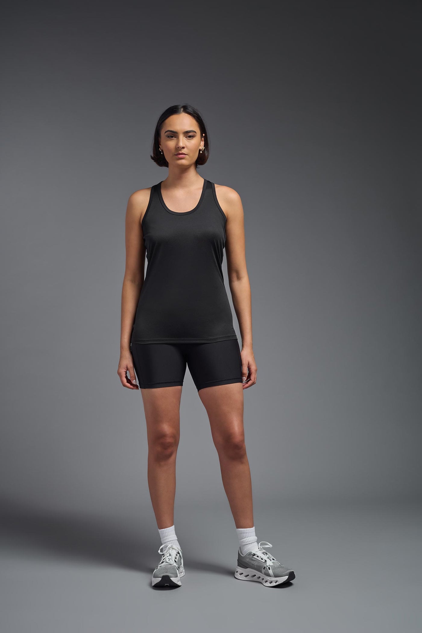 A female model with brown short hair on a gray background, wearing the Women's Drimax Endurance Racerback Tank in the color black from a full body front view. This tank is made in the USA and is quick-drying and moisture-wicking. #color_black