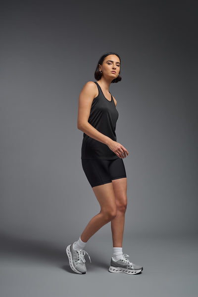 A female model with brown short hair on a gray background, wearing the Women's Drimax Endurance Racerback Tank in the color black from a full body side view. This tank is made in the USA and is quick-drying and moisture-wicking. #color_black