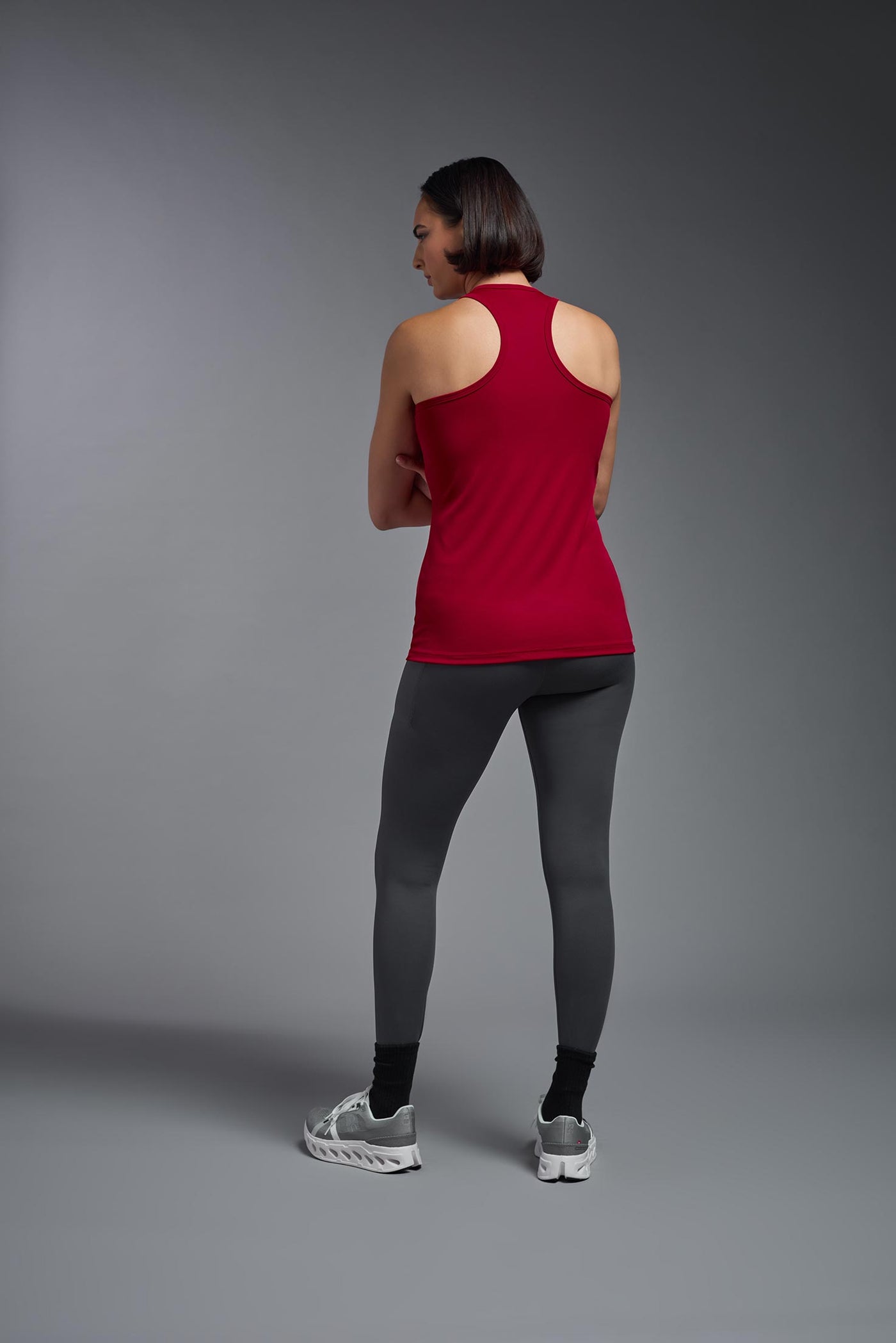 A female model with brown short hair on a gray background, wearing the Women's Drimax Endurance Racerback Tank in the color true red from a full body back view. This tank is made in the USA and is quick-drying and moisture-wicking. #color_true-red