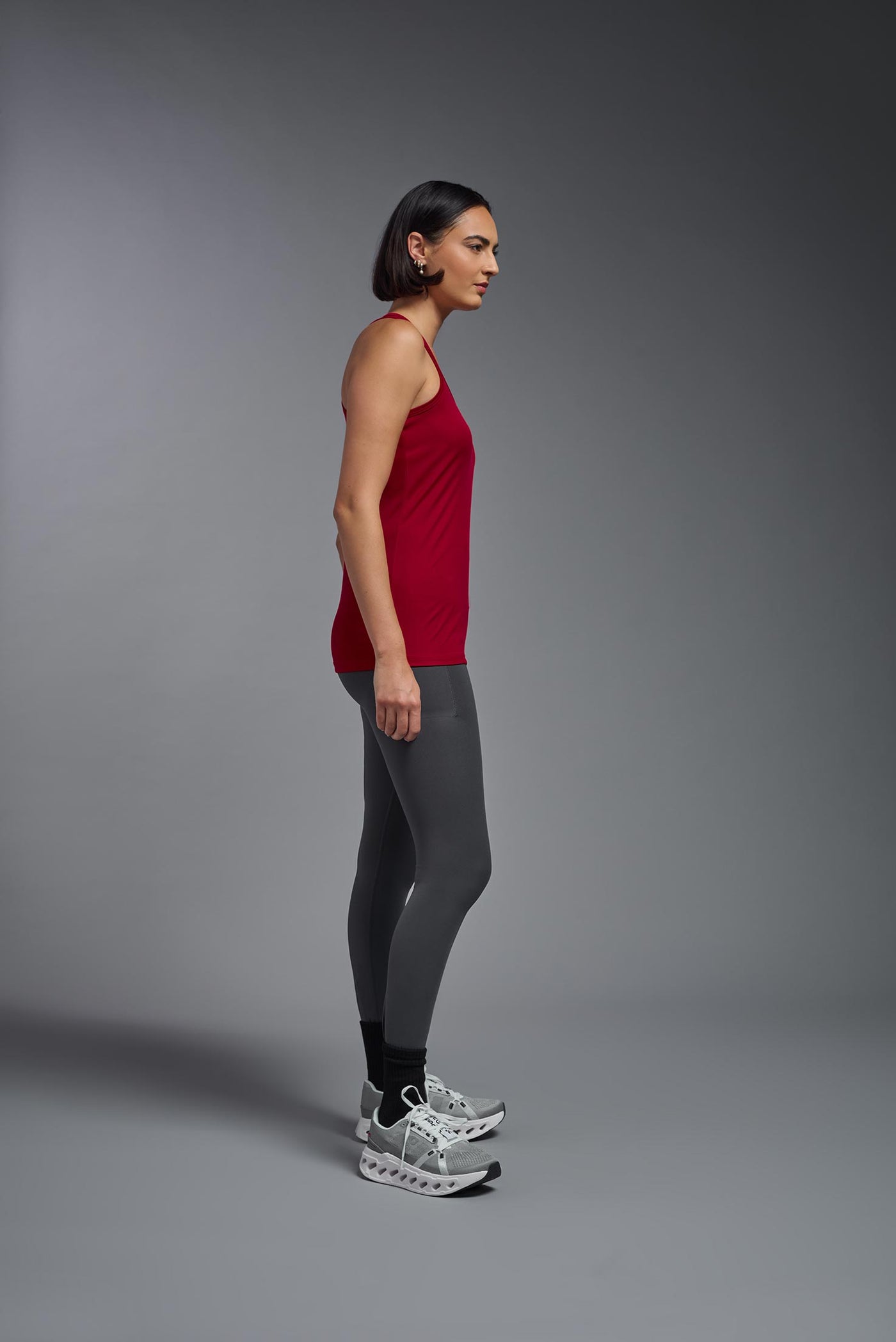 A female model with brown short hair on a gray background, wearing the Women's Drimax Endurance Racerback Tank in the color true red from a full body side view. This tank is made in the USA and is quick-drying and moisture-wicking. #color_true-red