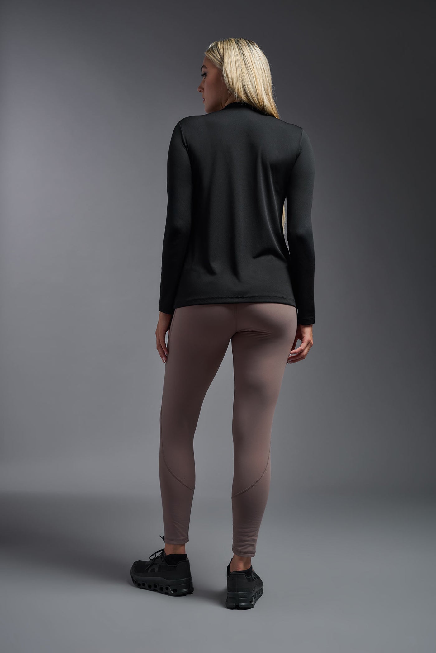 A blonde female model with long straight hair on a gray background, wearing the Women's DriMax™ V-neck Long Sleeve Expert Tee in the color black from a full body back view. This shirt is made in the USA and is quick-drying and moisture-wicking. #color_black