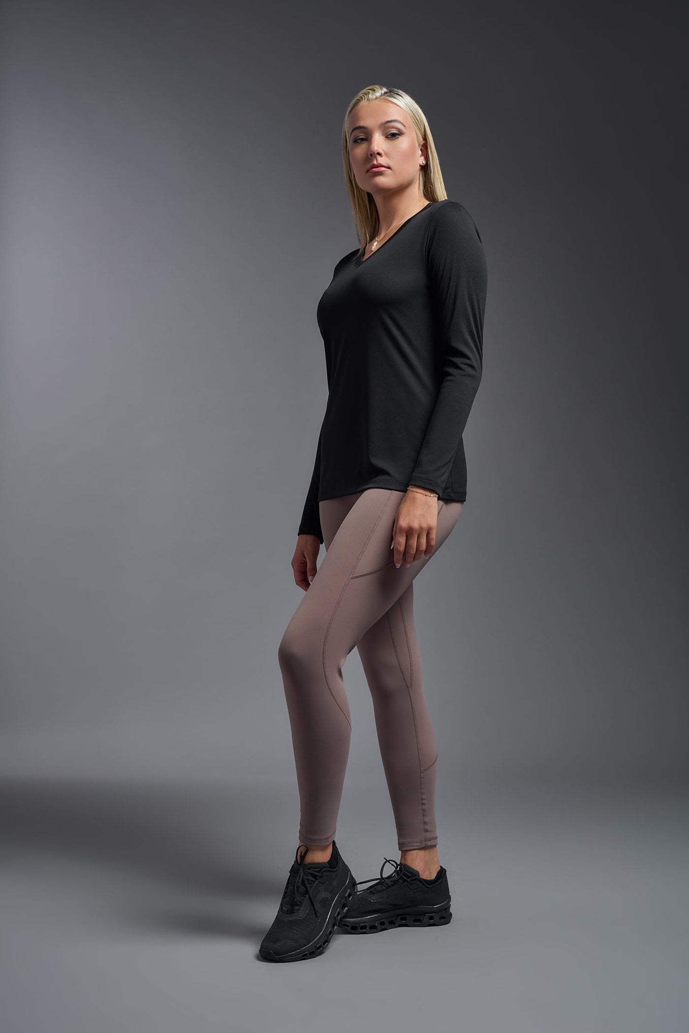 A blonde female model with long straight hair on a gray background, wearing the Women's DriMax™ V-neck Long Sleeve Expert Tee in the color black from a full body side view. This shirt is made in the USA and is quick-drying and moisture-wicking. #color_black
