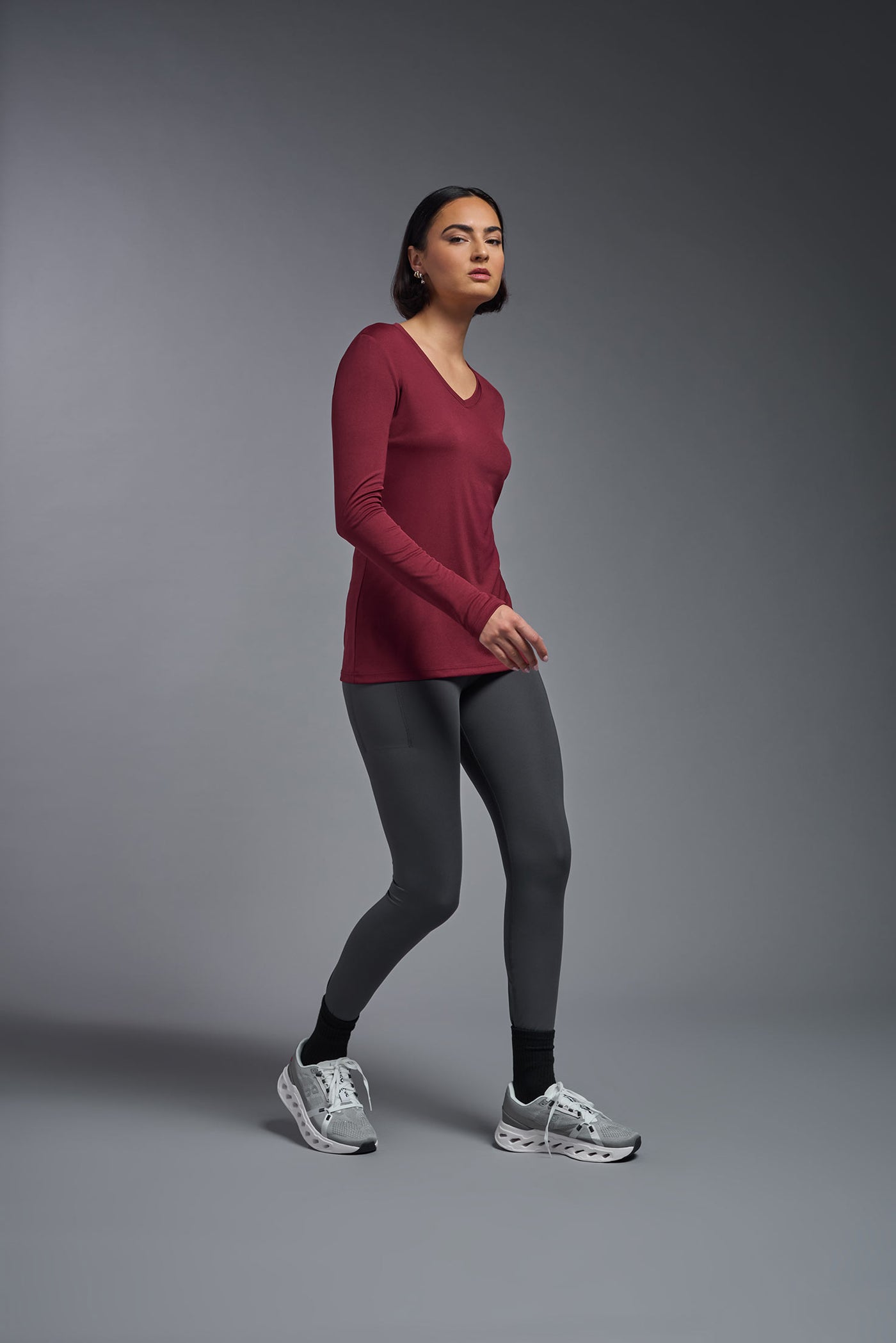 A female model with short brown hair on a gray background, wearing the Women's DriMax™ V-neck Long Sleeve Expert Tee in the color cardinal from a full body side view. This shirt is made in the USA and is quick-drying and moisture-wicking. #color_cardinal
