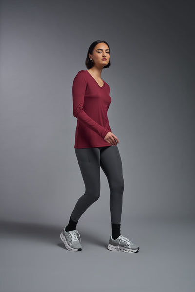 A female model with short brown hair on a gray background, wearing the Women's DriMax™ V-neck Long Sleeve Expert Tee in the color cardinal from a full body side view. This shirt is made in the USA and is quick-drying and moisture-wicking. #color_cardinal
