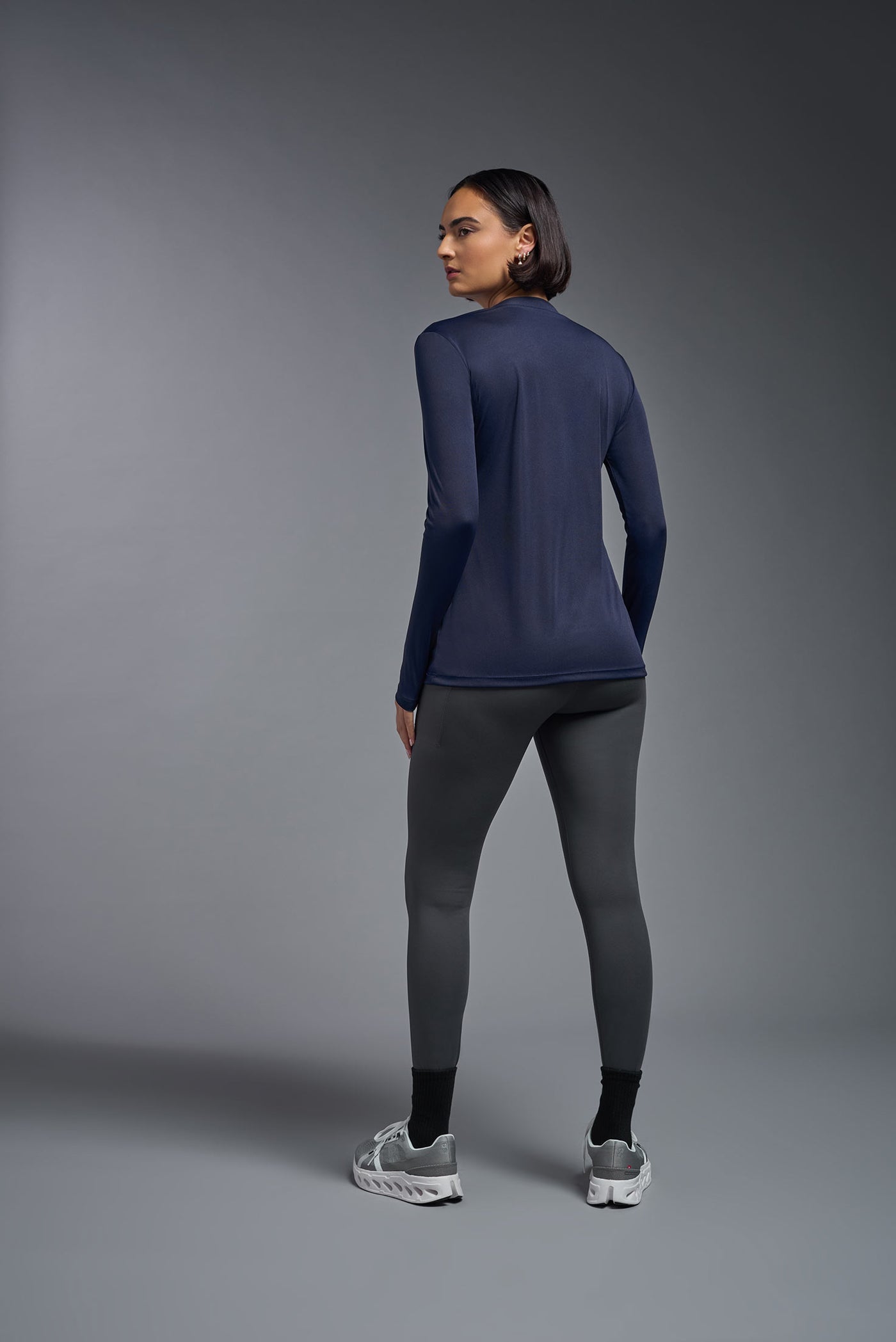 A female model with short brown hair on a gray background, wearing the Women's DriMax™ V-neck Long Sleeve Expert Tee in the color navy from a closeup collar back view. This shirt is made in the USA and is quick-drying and moisture-wicking. #color_navy