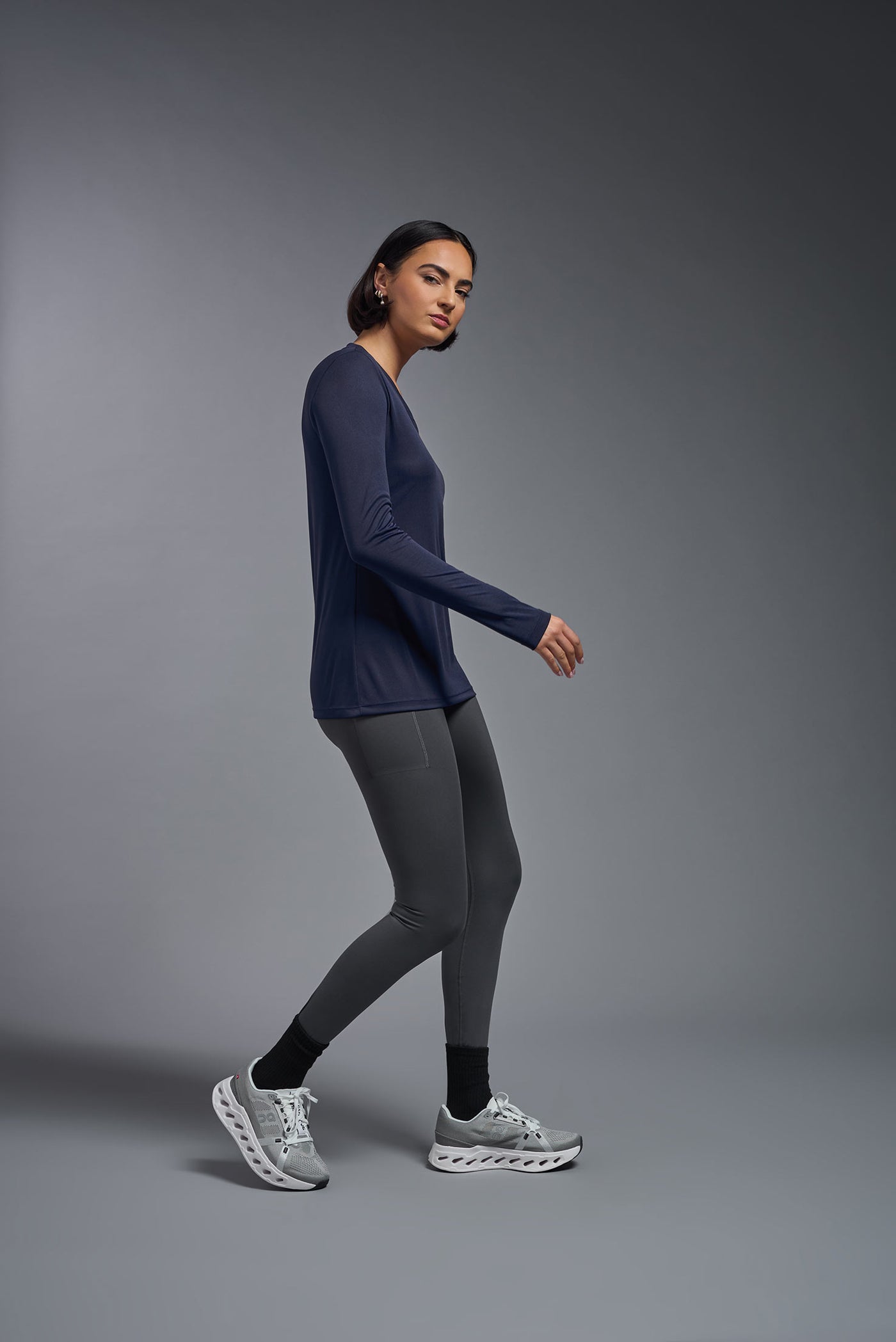 A female model with short brown hair on a gray background, wearing the Women's DriMax™ V-neck Long Sleeve Expert Tee in the color navy from a full body side view. This shirt is made in the USA and is quick-drying and moisture-wicking. #color_navy