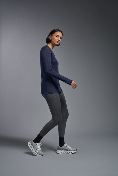 A female model with short brown hair on a gray background, wearing the Women's DriMax™ V-neck Long Sleeve Expert Tee in the color navy from a full body side view. This shirt is made in the USA and is quick-drying and moisture-wicking. #color_navy