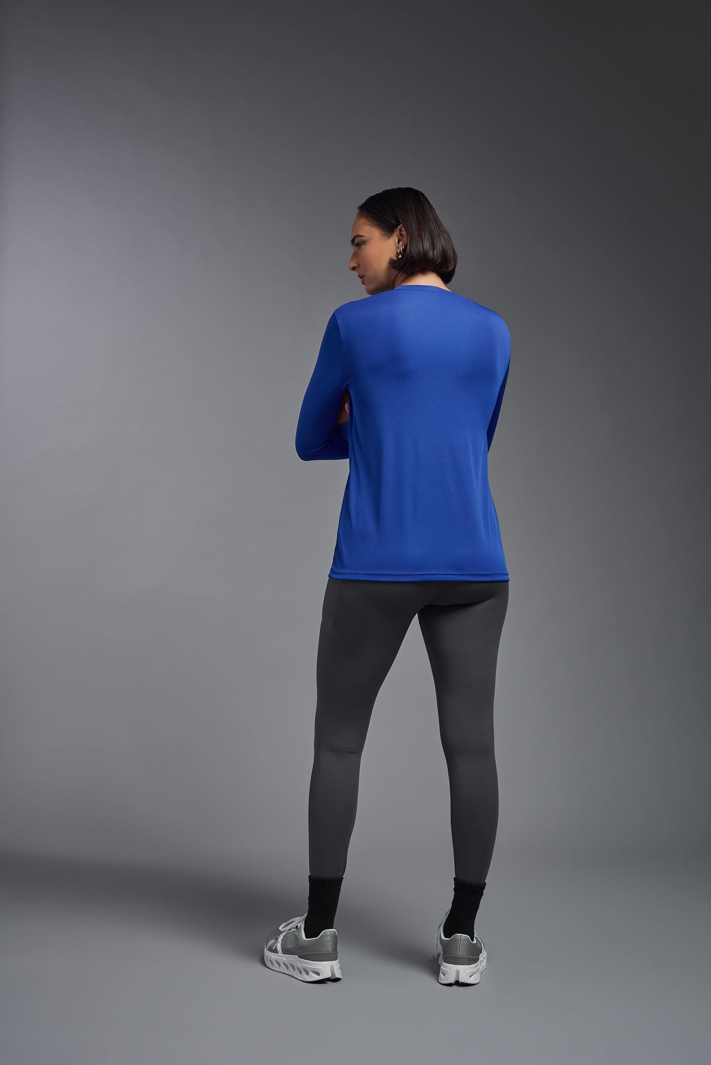 A female model with short brown hair on a gray background, wearing the Women's DriMax™ V-neck Long Sleeve Expert Tee in the color royal blue from a full body back view. This shirt is made in the USA and is quick-drying and moisture-wicking. #color_royal-blue