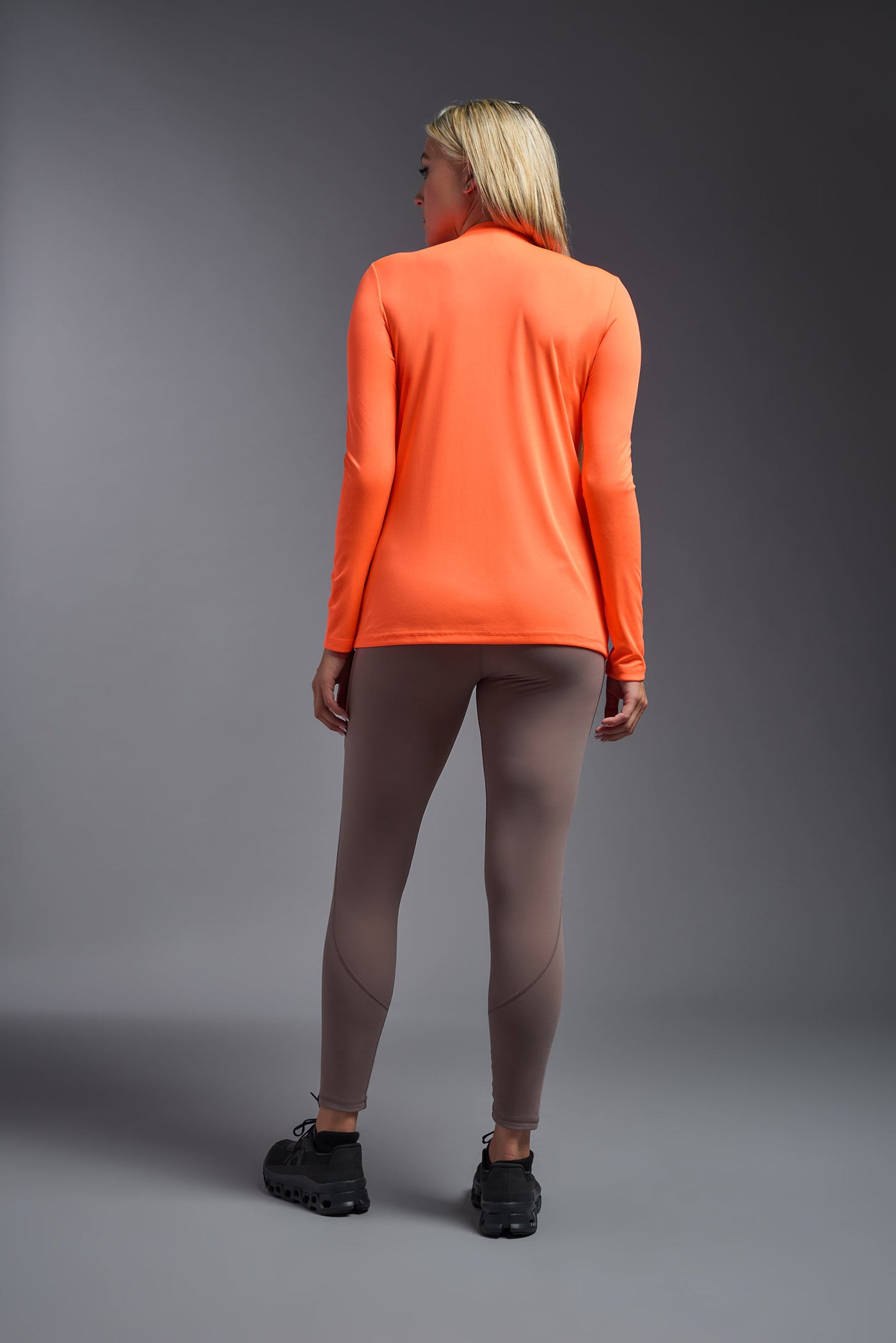 A female model with short brown hair on a gray background, wearing the Women's DriMax™ V-neck Long Sleeve Expert Tee in the color safety orange from a full body back view. This shirt is made in the USA and is quick-drying and moisture-wicking. #color_safety-orange